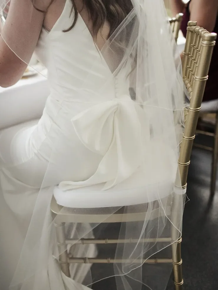 Close-up of a bride wearing a white wedding gown with a large bow at the waist, sitting on a gold-colored chair with a sheer veil flowing down from her head.