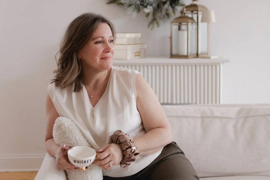 Wedding Planner sitting on a couch holding a whiskey glass and a white towel, looking out thoughtfully in a cozy, decorated living room.