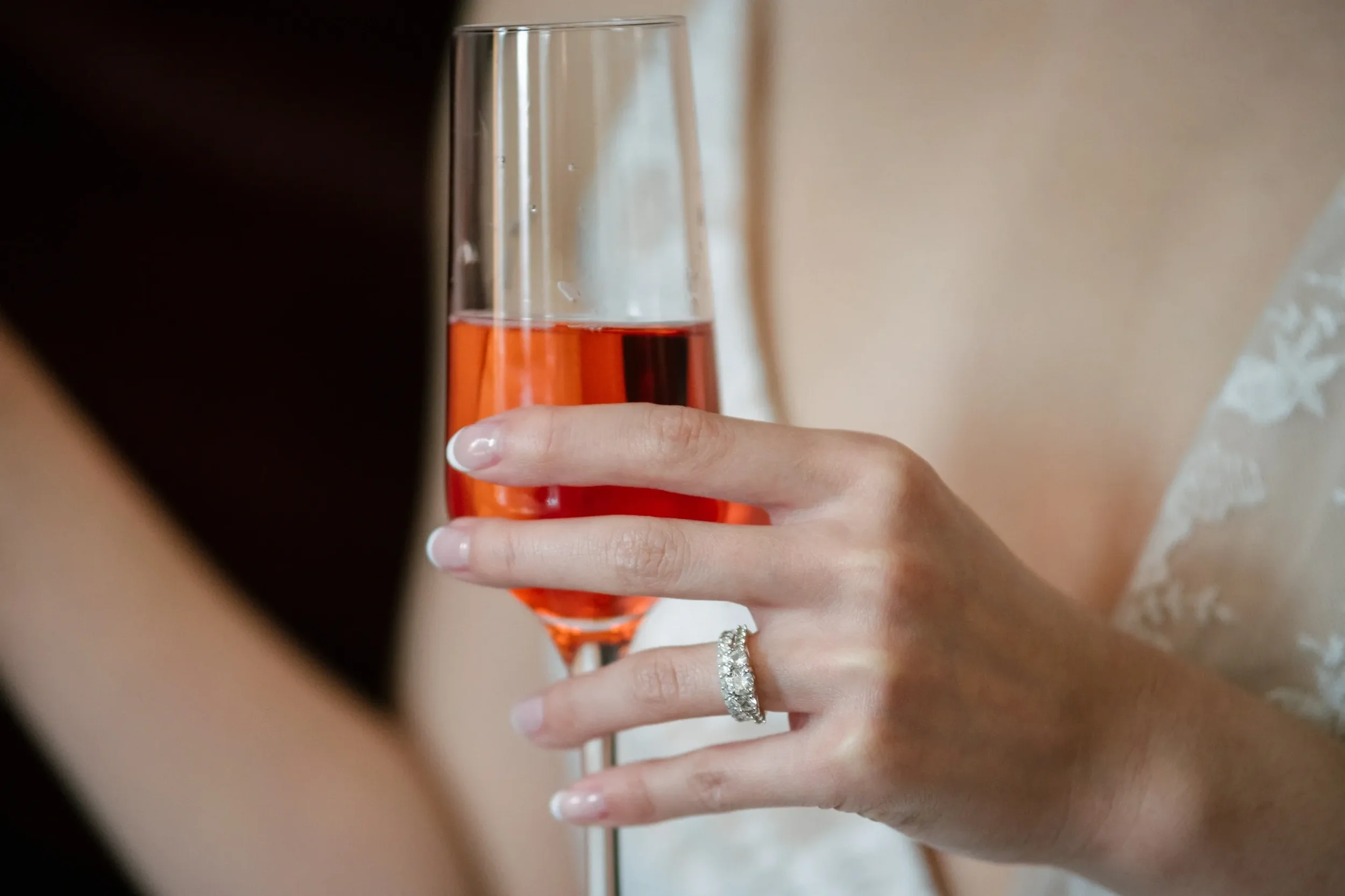 A woman holding a glass of pink sparkling wine, wearing a wedding dress and a diamond ring.