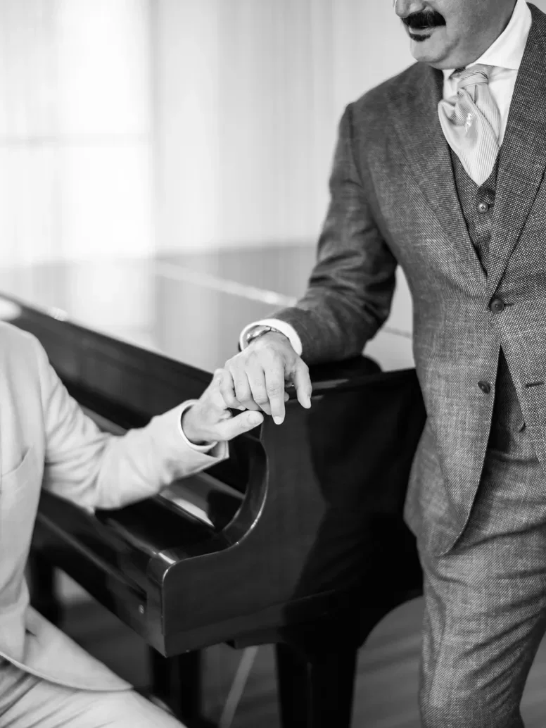 Grooms dressed in a suits gently holding hands with each other at a piano.