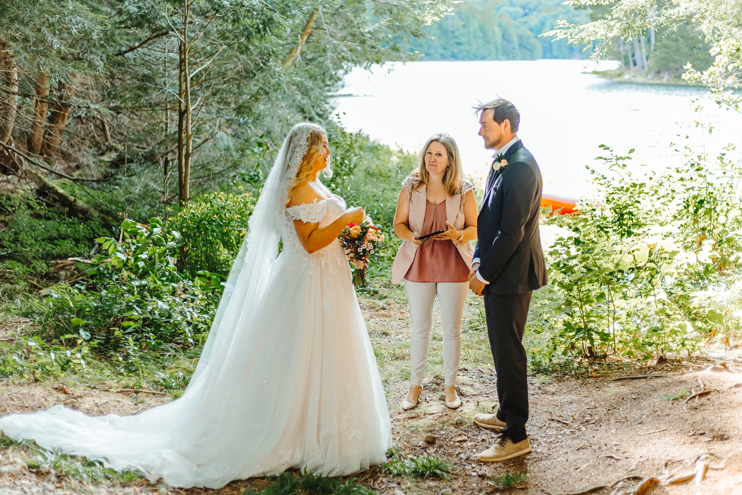 A bride and groom standing in an outdoor wooded area by a river, with an officiant holding a tablet or laptop between them.