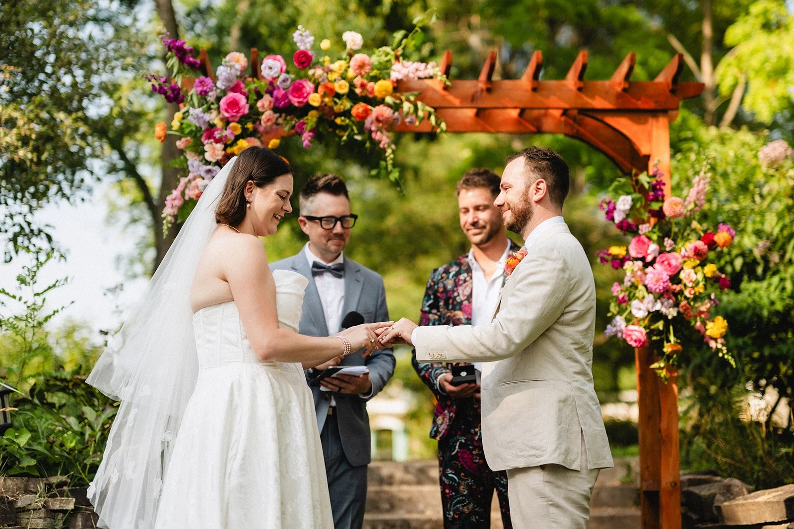 A couple getting married outdoors under a floral arch, with officiant and ring bearer present, on a sunny day with greenery background.