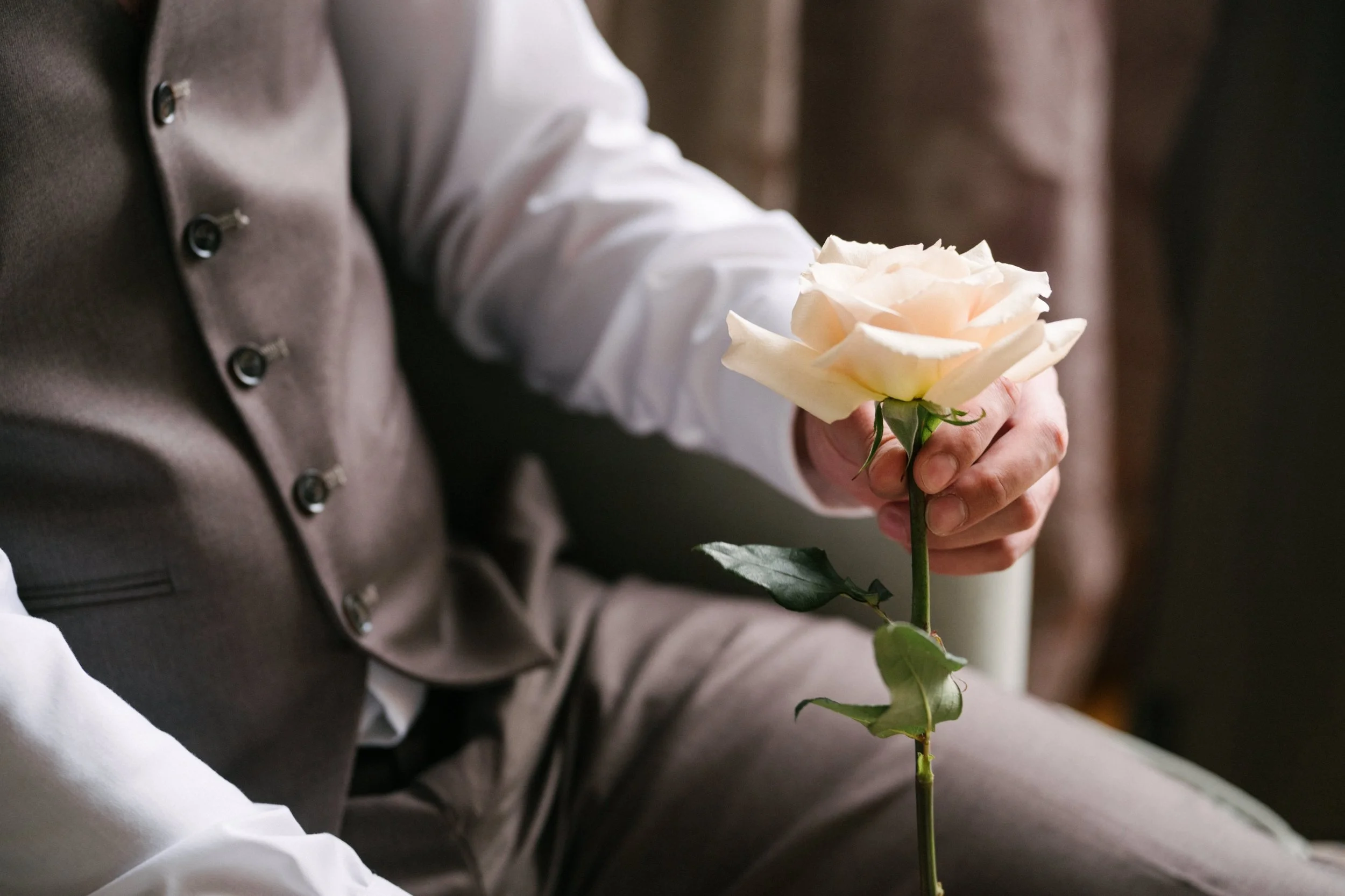 Groom wearing a brown vest and white shirt holding a pale pink rose with green leaves.