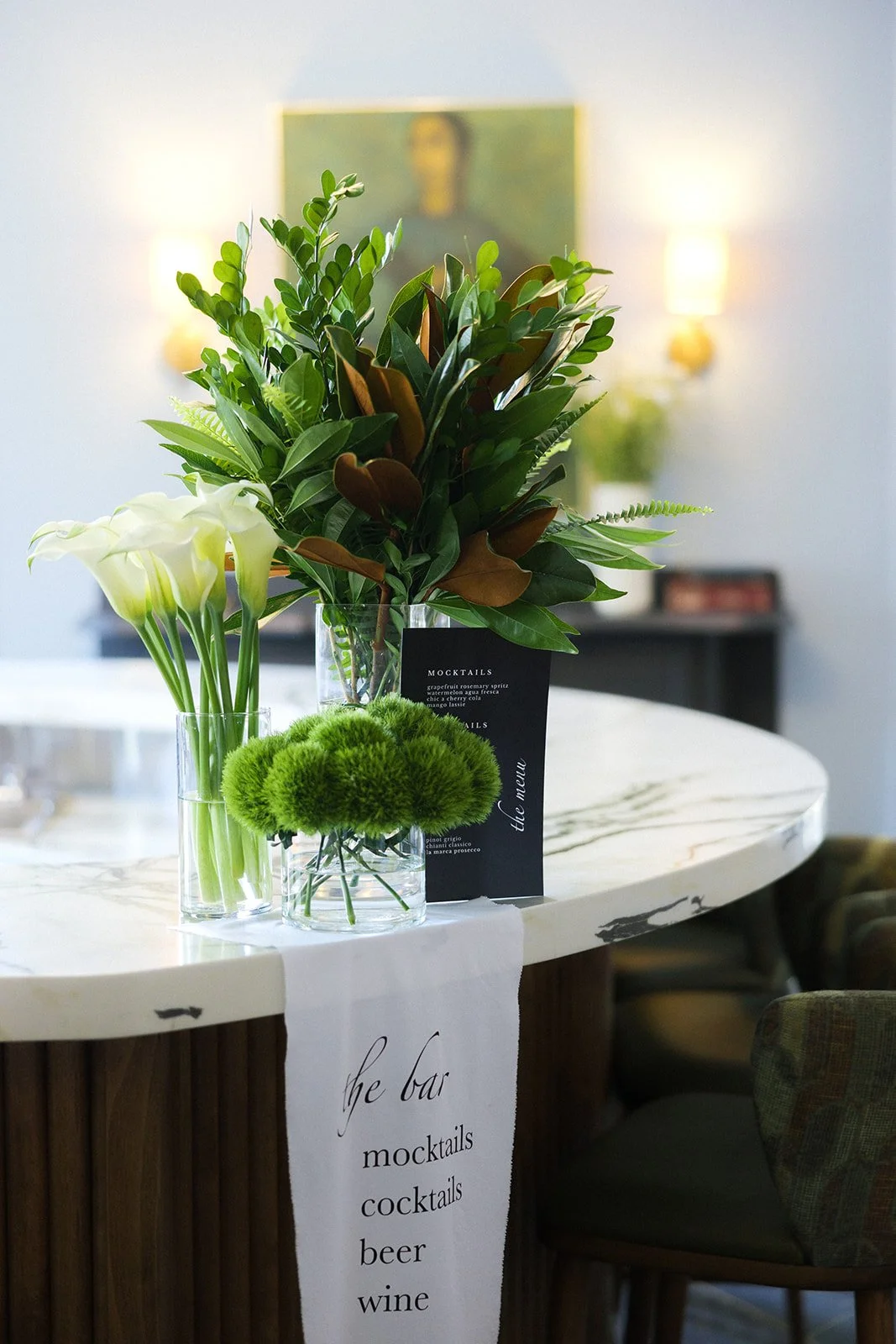 Decorative floral arrangement on a bar counter, featuring white calla lilies, green flowers, and lush green foliage, with a white cloth hanging from the counter that reads 'the bar, mocktails, cocktails, beer, wine'.