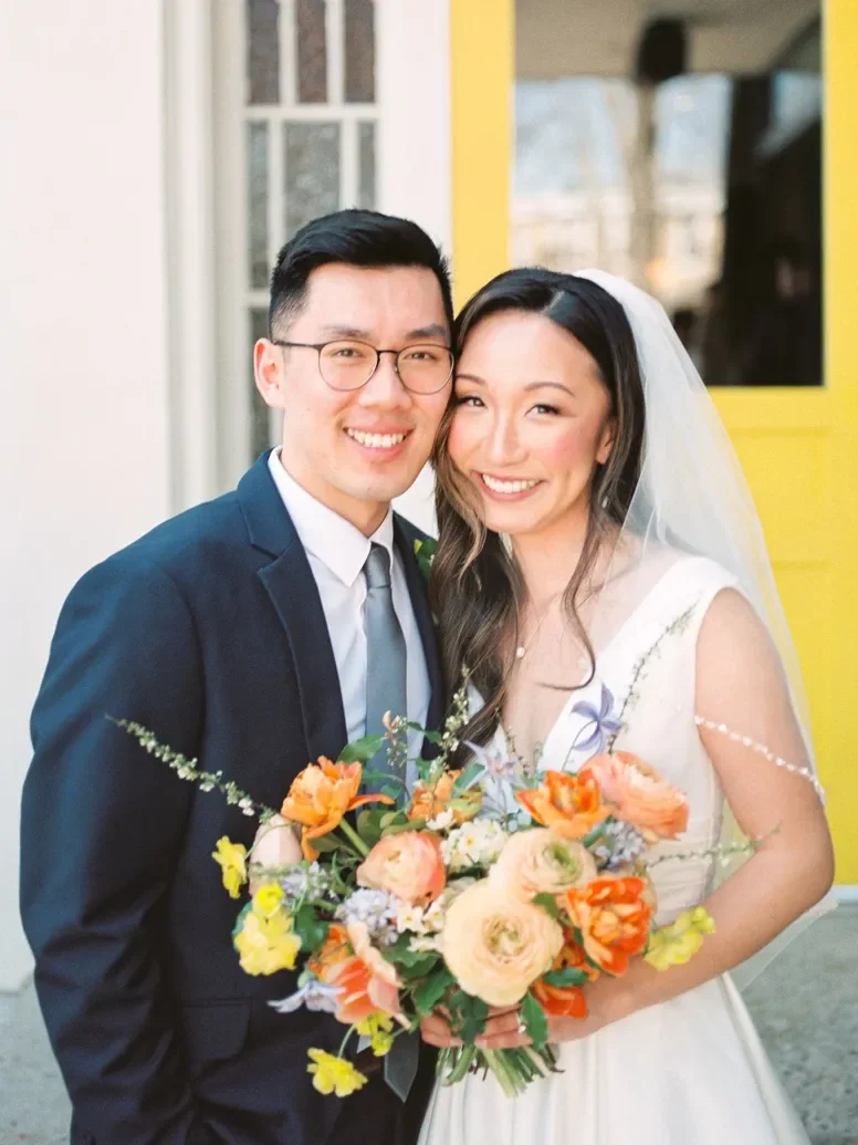 A bride and groom smiling and posing for a photo, with the bride holding a colorful bouquet. The groom is wearing a dark suit and glasses, and the bride is in a white wedding dress with a veil.