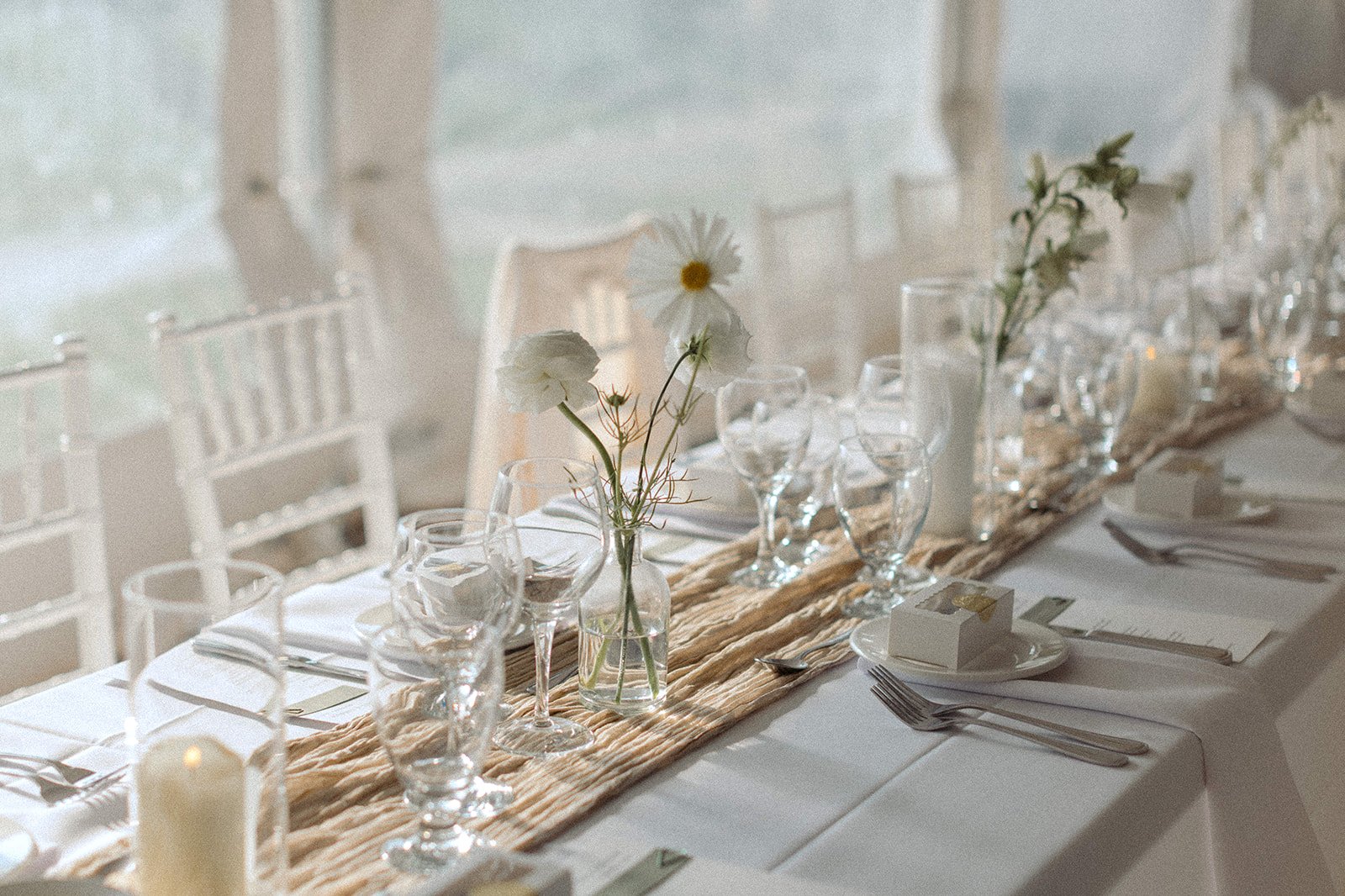 Elegant dining table setup with white tablecloth, glassware, silverware, and small floral centerpieces in glass vases, decorated with a beige table runner and candles, in a softly lit room with large windows.