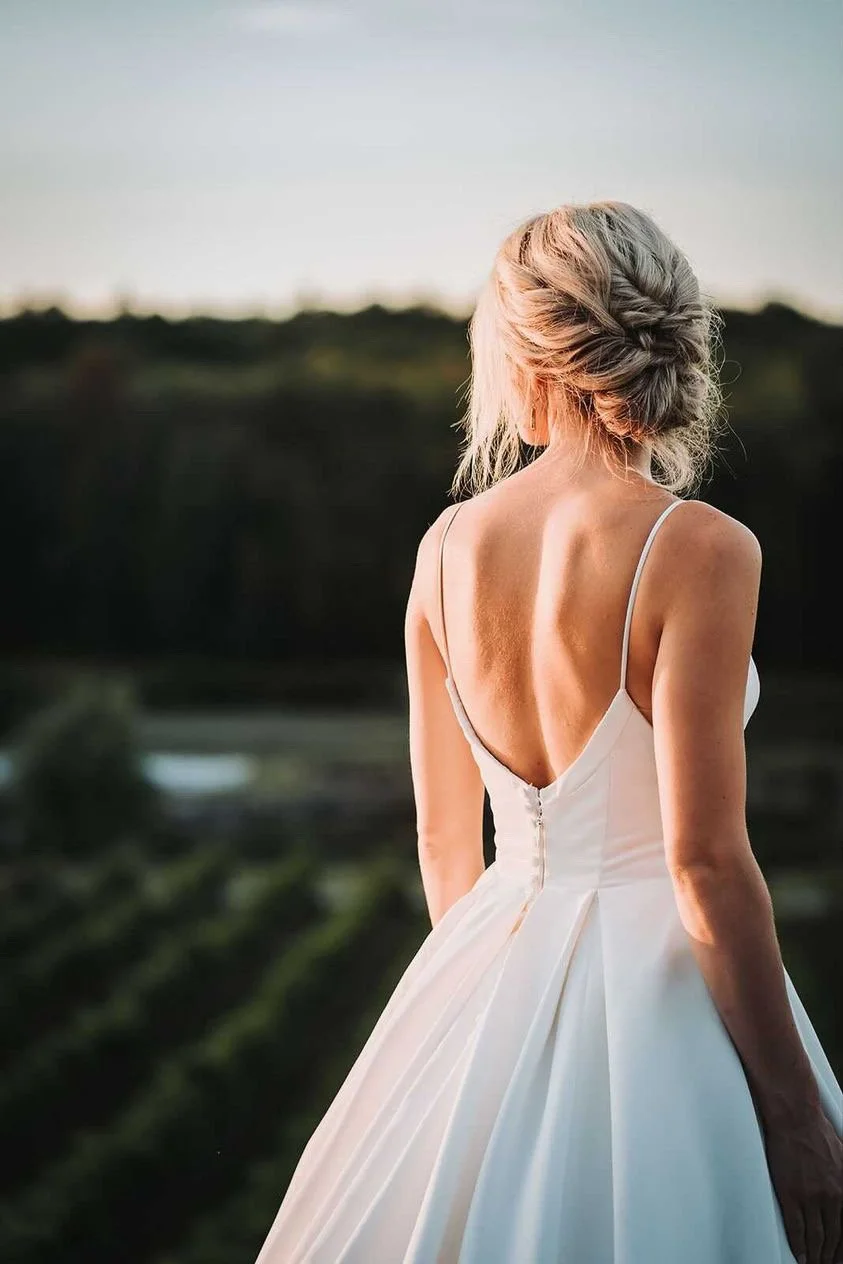 A bride with blonde hair styled in an elegant updo is wearing a white wedding dress with thin straps, standing outdoors during sunset.