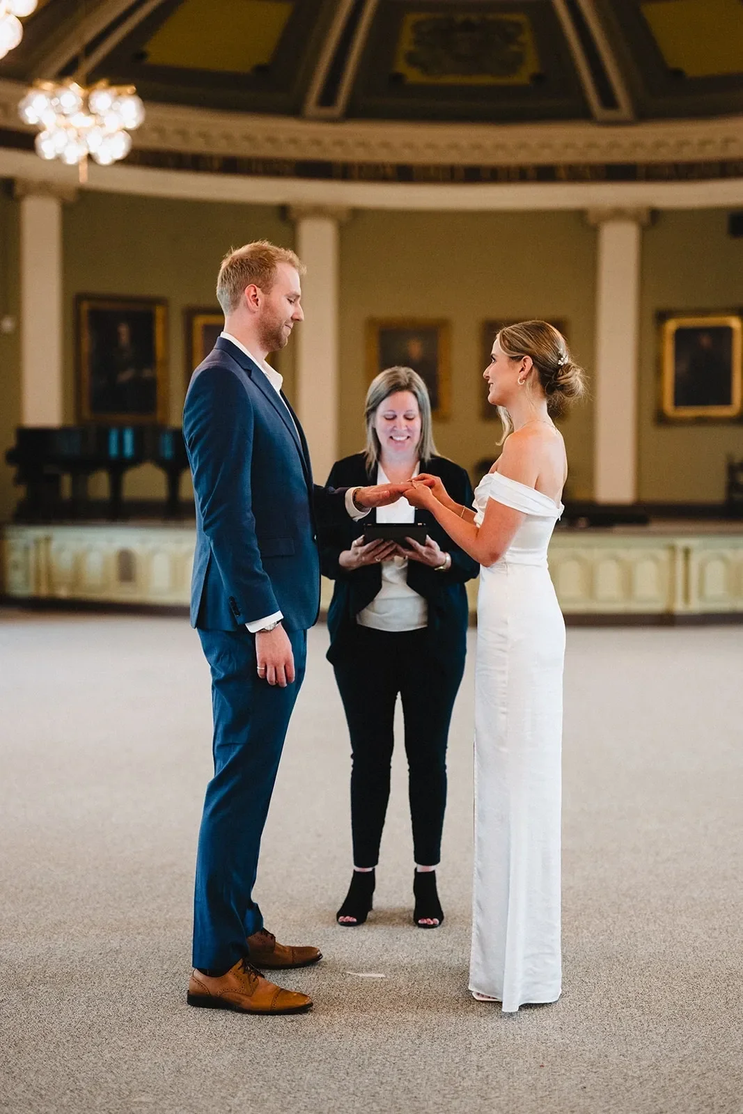 A couple getting married in a formal indoor setting, with the bride placing a ring on the groom's finger while a woman officiant watches and smiles.