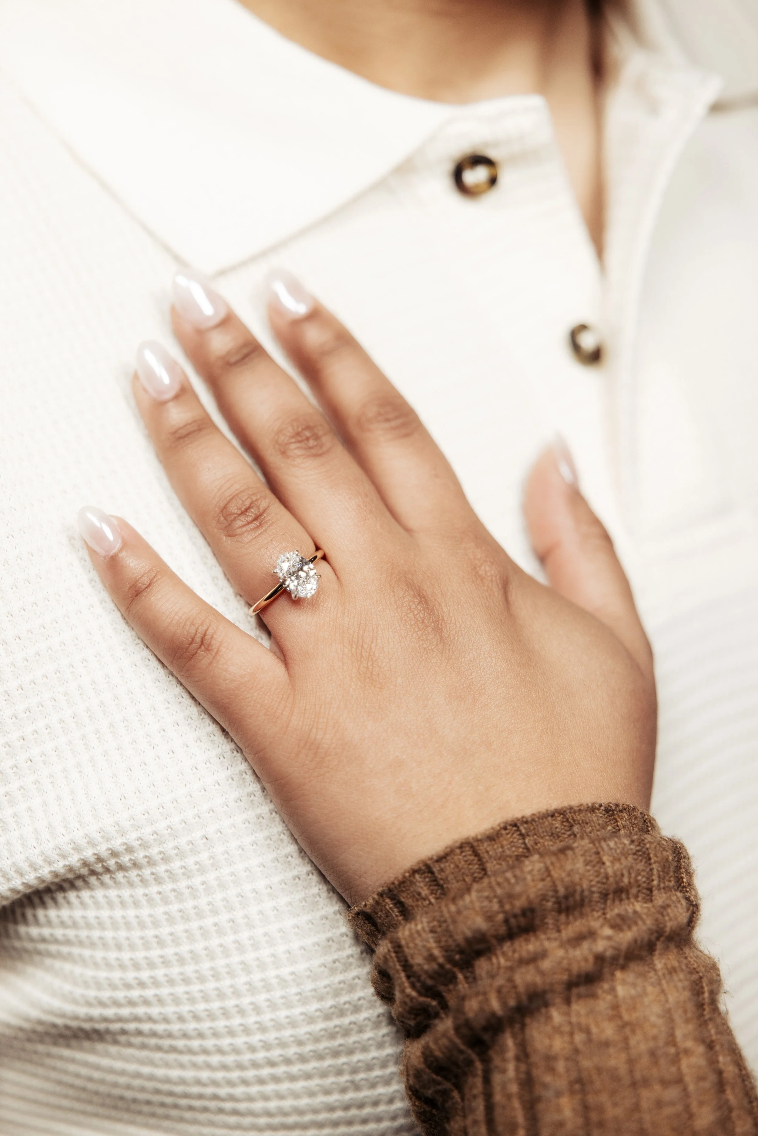 A close-up of a woman's hand wearing a diamond engagement ring on her ring finger, resting on her chest, with a beige and white outfit.