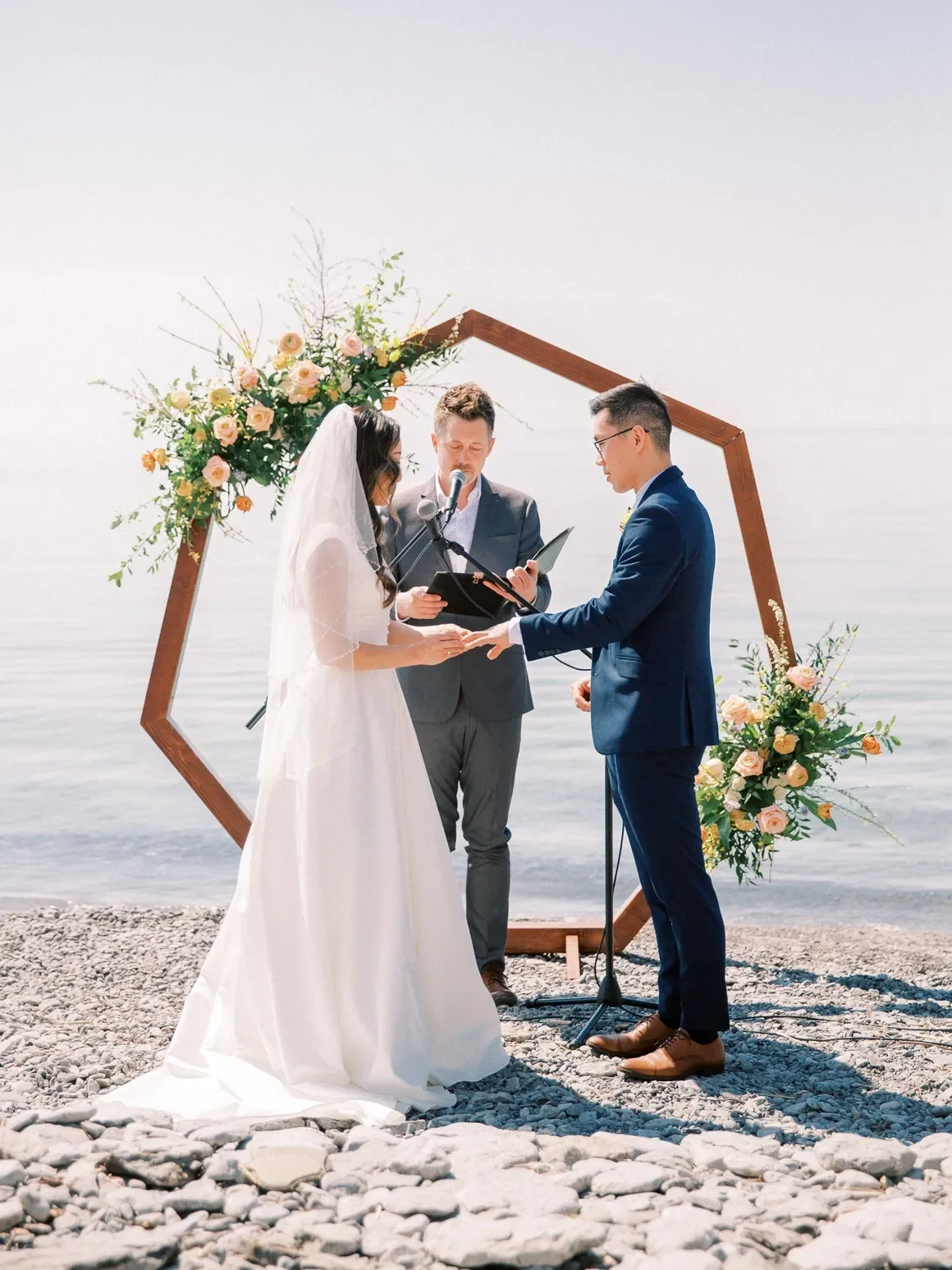 Bride and groom with officiant standing infront of a hexagon arch with florals on a rocky beach at Lake Ontario