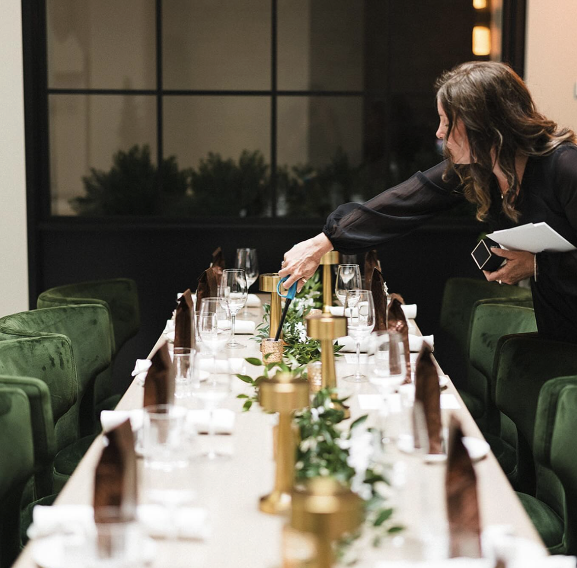 Wedding planner arranging a decorated dining table with green chairs, gold candle holders, and greenery, in a well-lit room with large windows.