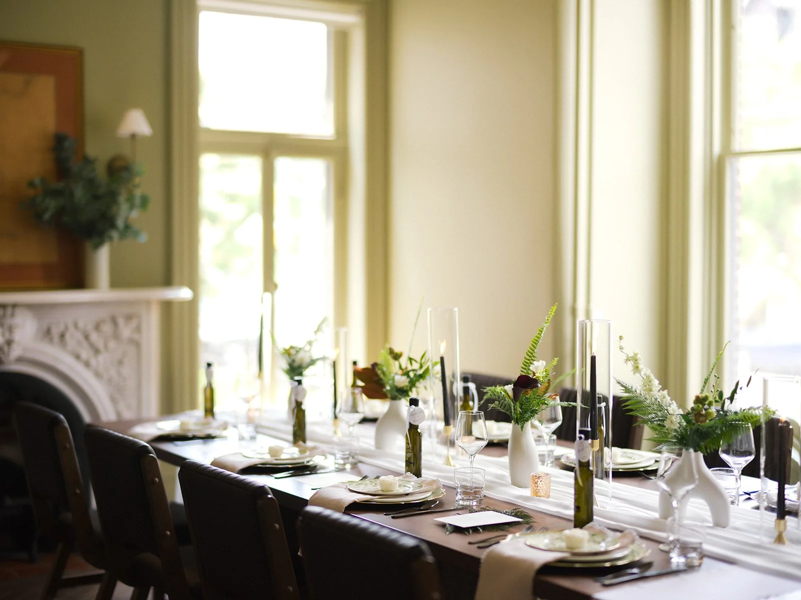 A long dining table set for a formal event with flowers, candles, and dinnerware, in a bright room with large windows and natural light.