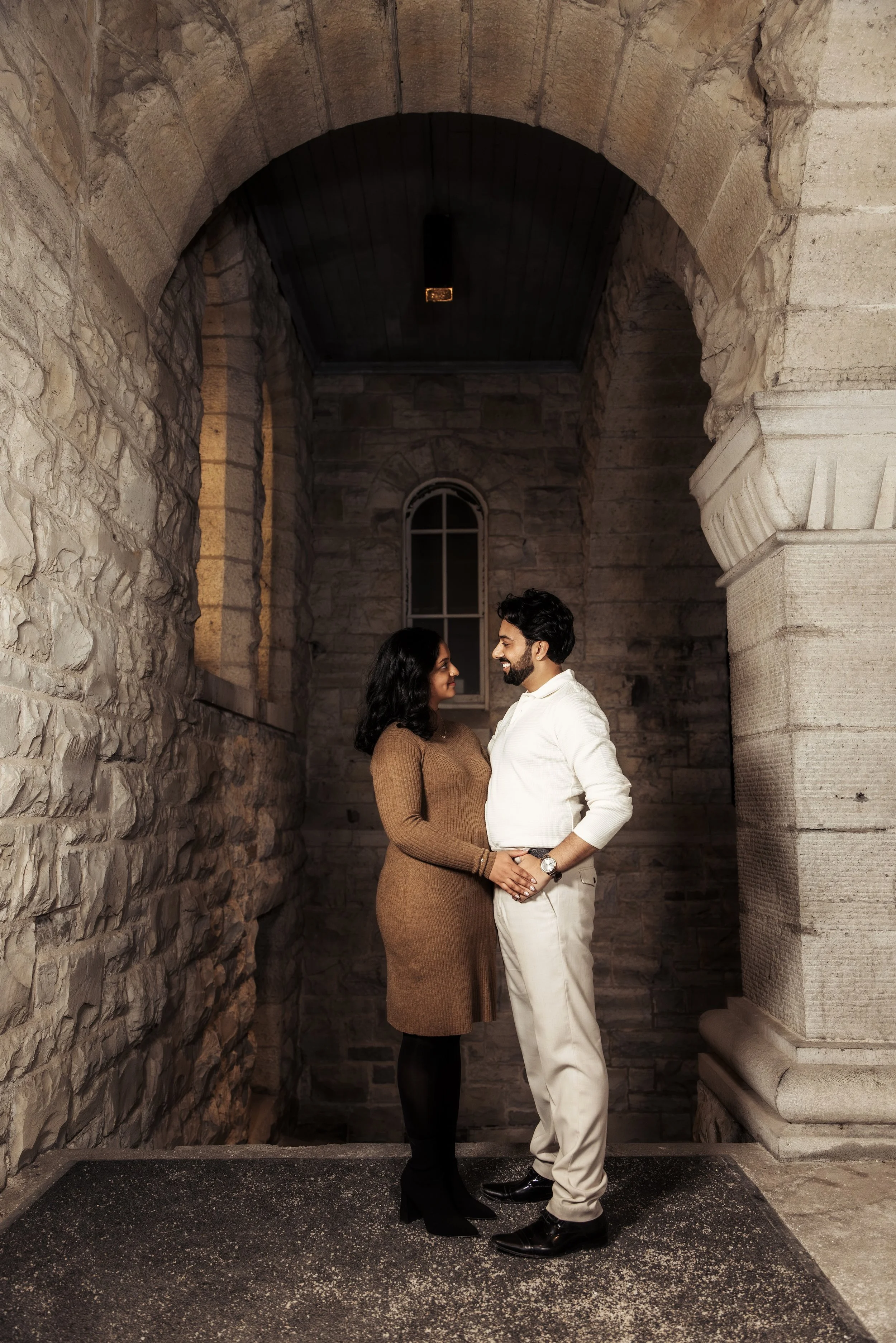 A couple stands facing each other in a stone archway, holding hands and smiling at each other indoors.