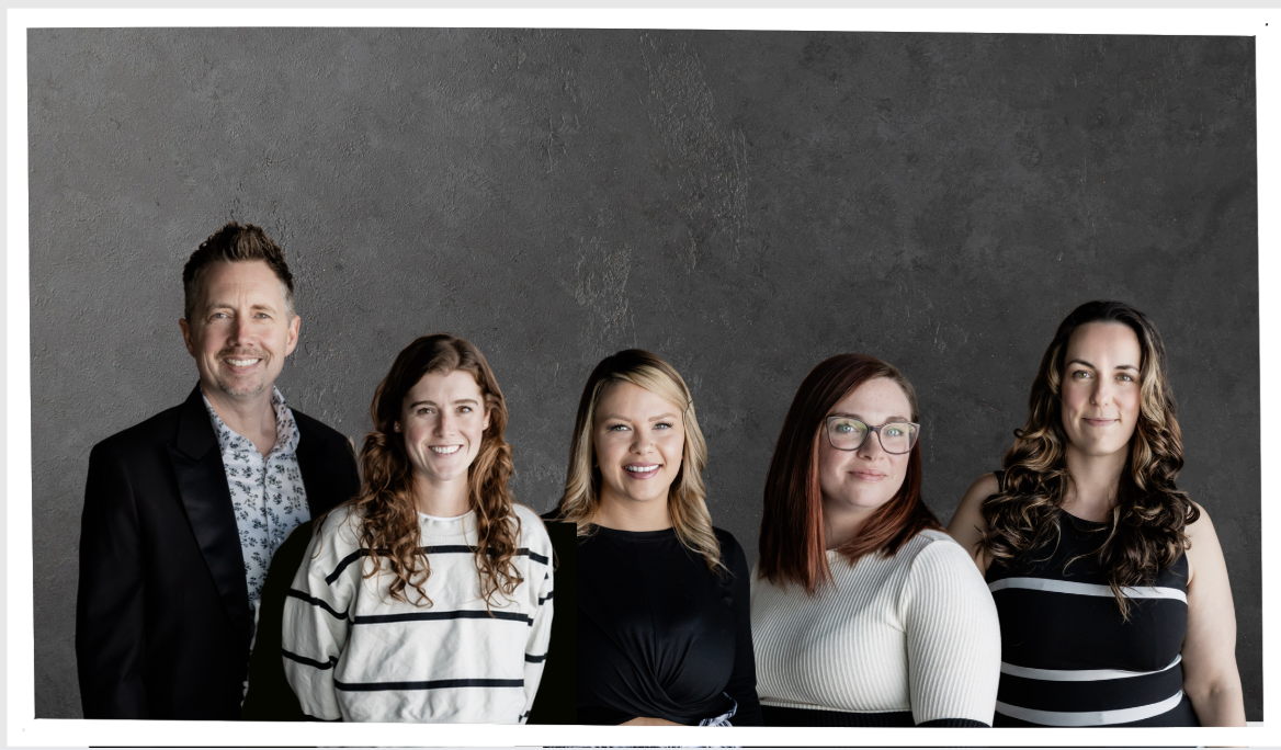 Group of five diverse adults standing against a textured gray wall, smiling.