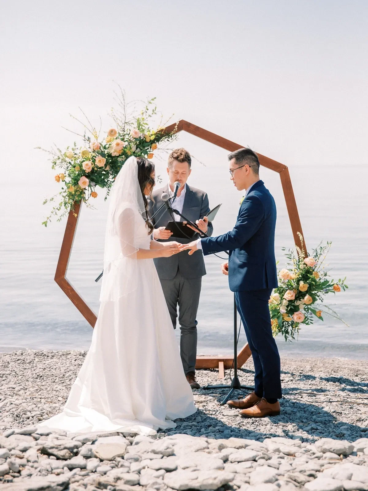 A couple getting married on a rocky beach with an ocean background, standing under a hexagonal wooden arch decorated with pink and peach flowers, with an officiant reading from a book.