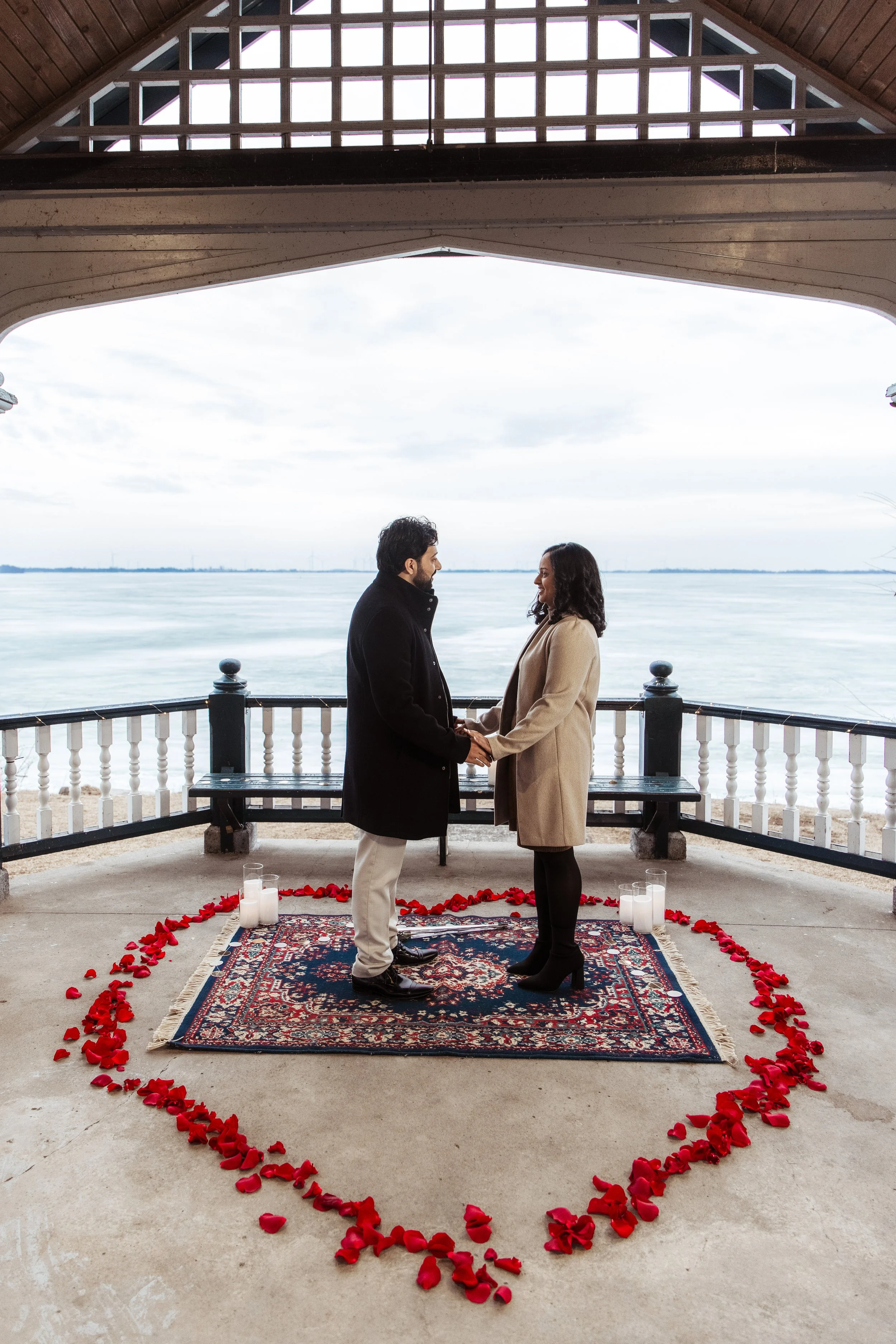 A man and woman holding hands, standing on a Persian rug with candles and rose petals arranged in a heart shape around them. They are in an outdoor covered area overlooking a body of water, with cloudy sky and wind turbines in the background. They appear to be having a romantic or wedding ceremony.
