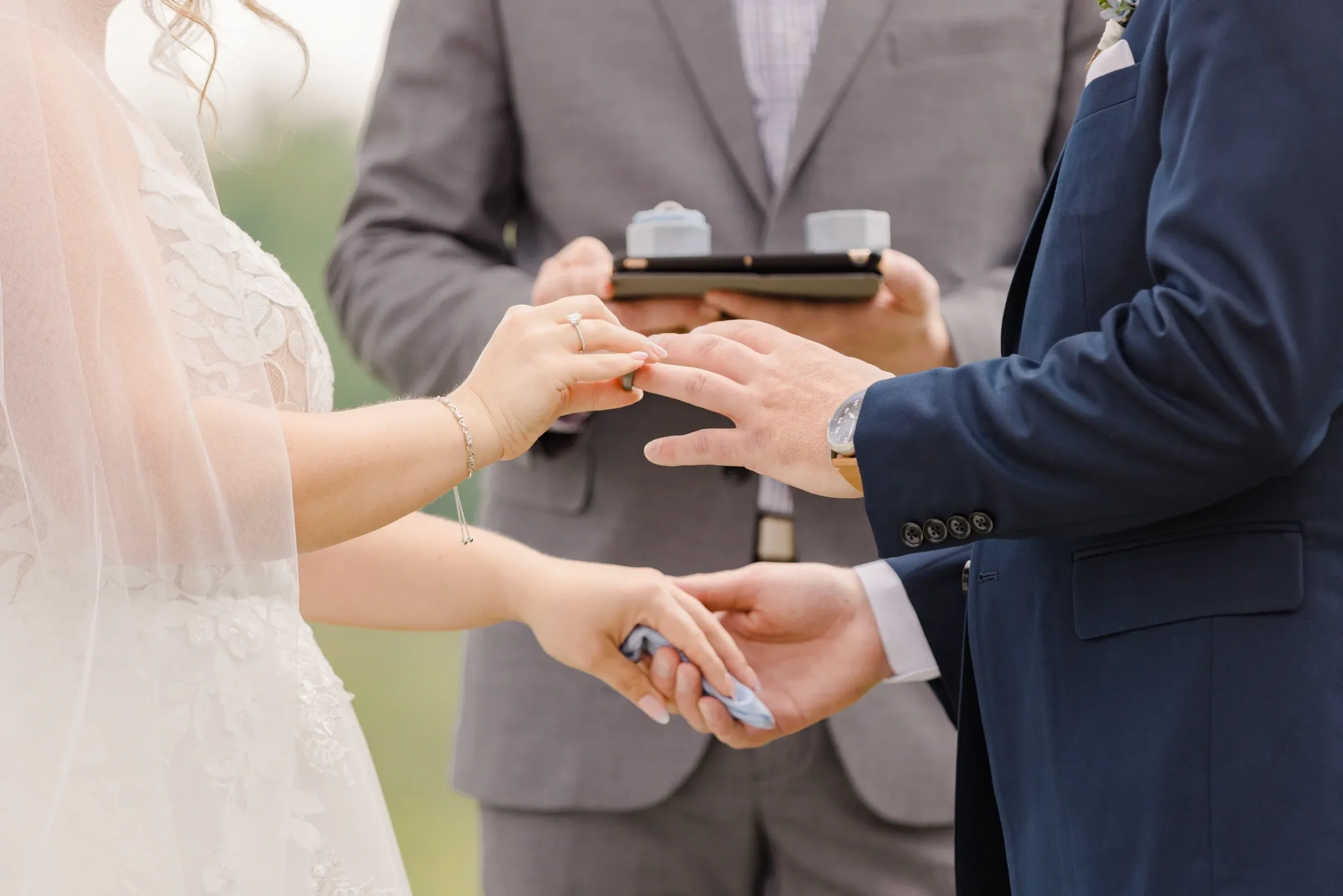 A bride and groom exchanging wedding rings during their ceremony, with a officiant holding a tablet in the background.