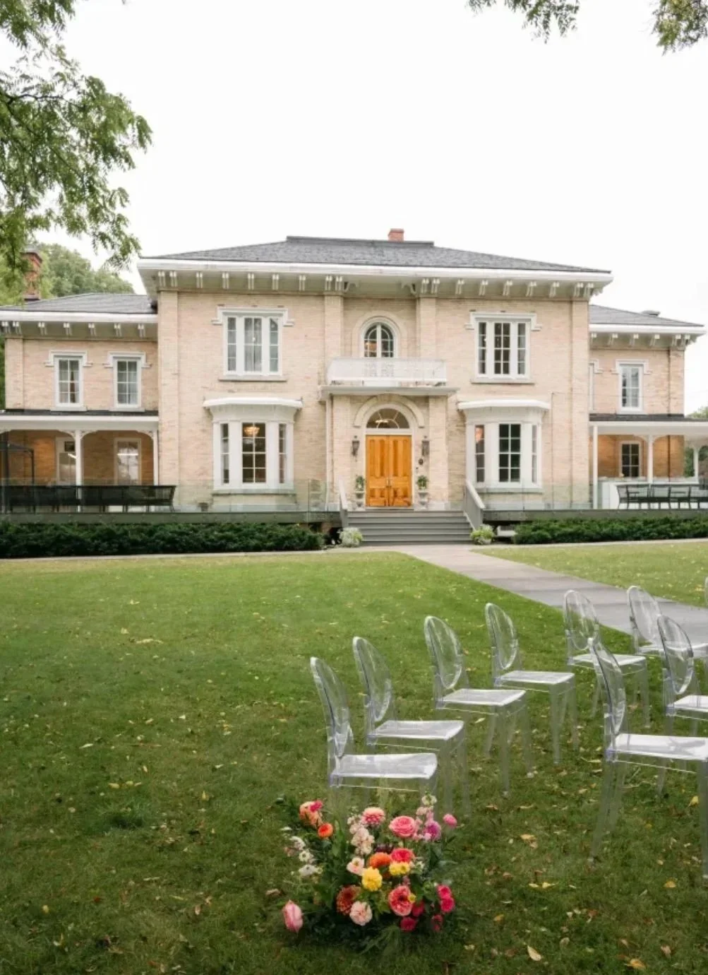 Stunning historic beige brick mansion with double front doors and a large wrap around porch. On the lawn are ghost chairs and flower set up for a ceremony.