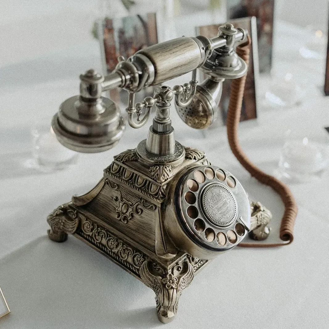 An antique metallic rotary telephone with ornate engravings on the base, placed on a white surface.