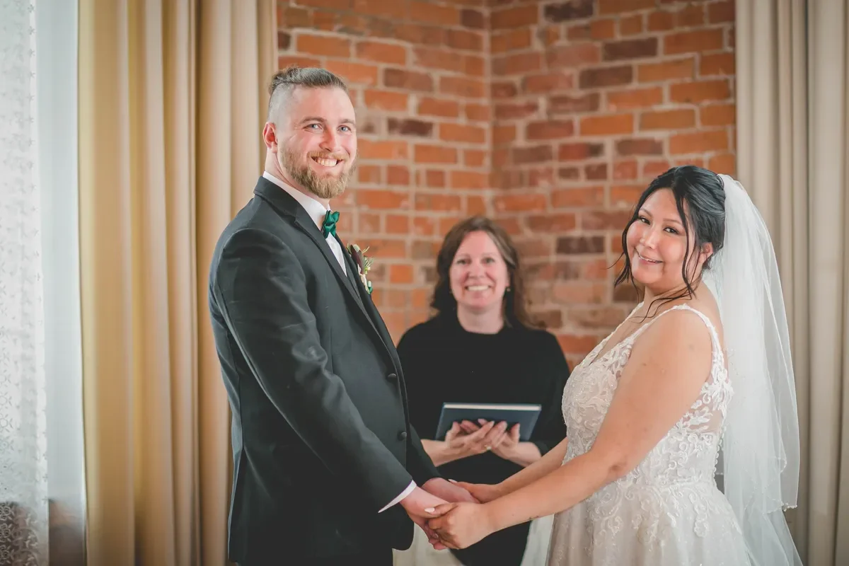 Bride and groom holding hands, smiling at each other, during their wedding ceremony. Officiant standing behind them, smiling.