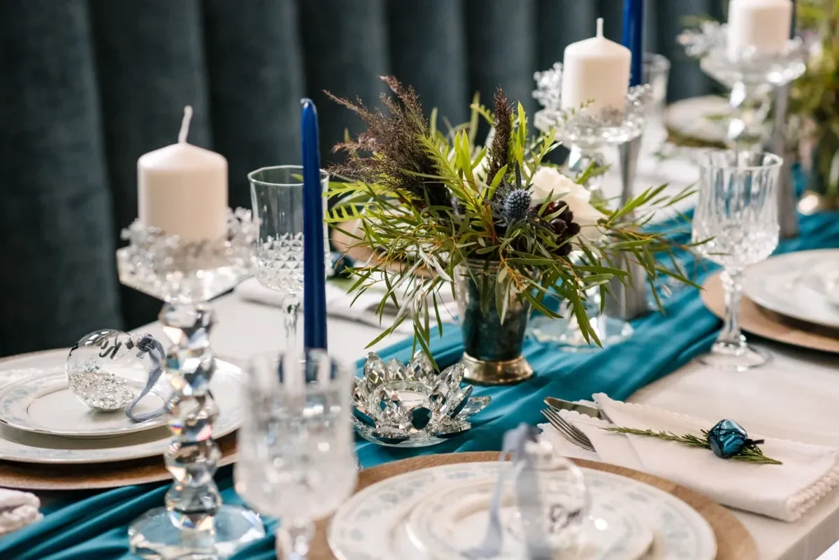 Elegant table setting with white plates, crystal glasses, and a centerpiece of flowers and greenery. White candles in glass holders and a teal table runner add decoration.