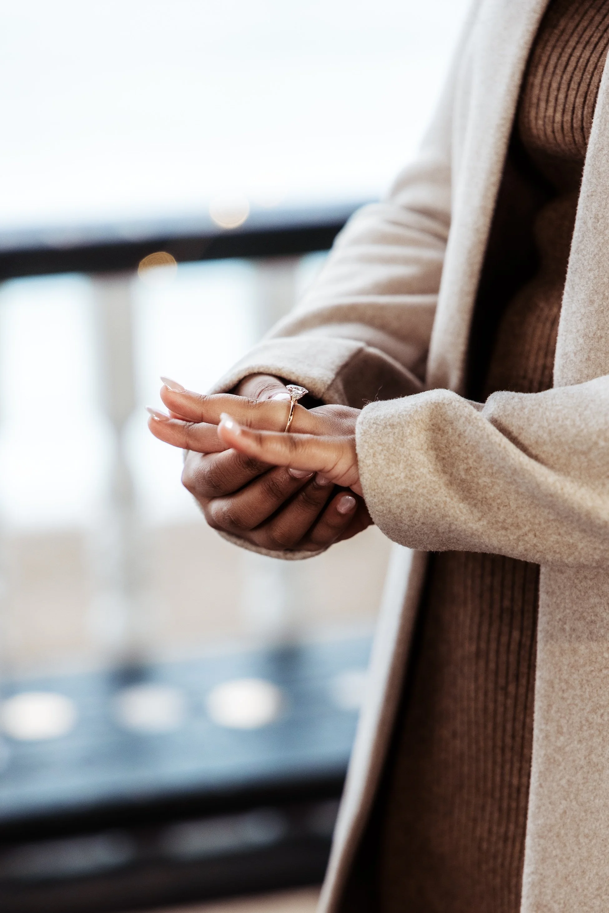 Close-up of a woman’s hands with a ring on her finger, wearing a beige coat and brown sweater, standing indoors near a window.