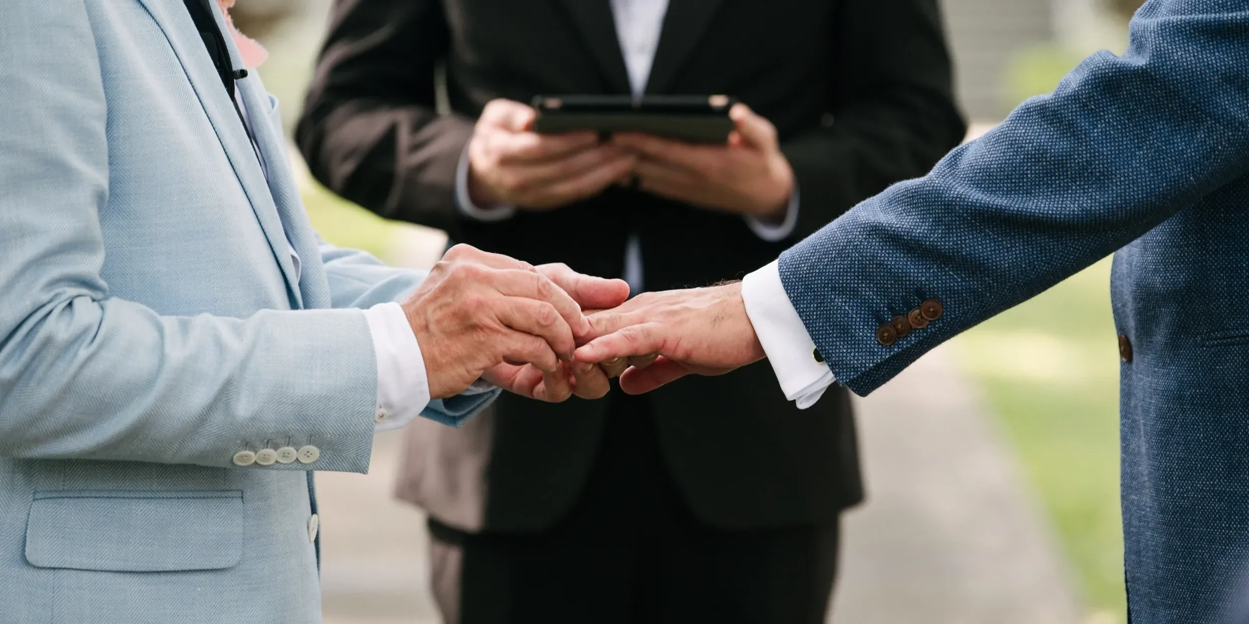 Grooms holding hands and one officiant in the background performing a wedding ceremony at the CAPE in Picton.