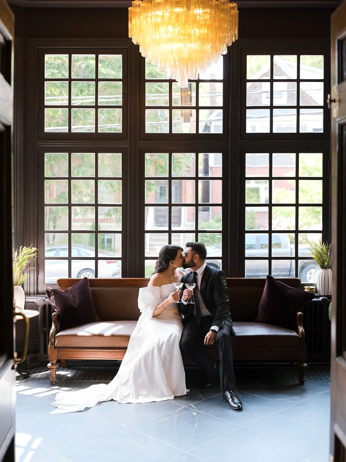 A bride and groom sitting on a vintage sofa, sharing a kiss and toasting with champagne glasses inside a bright room with large window panels and a chandelier overhead.