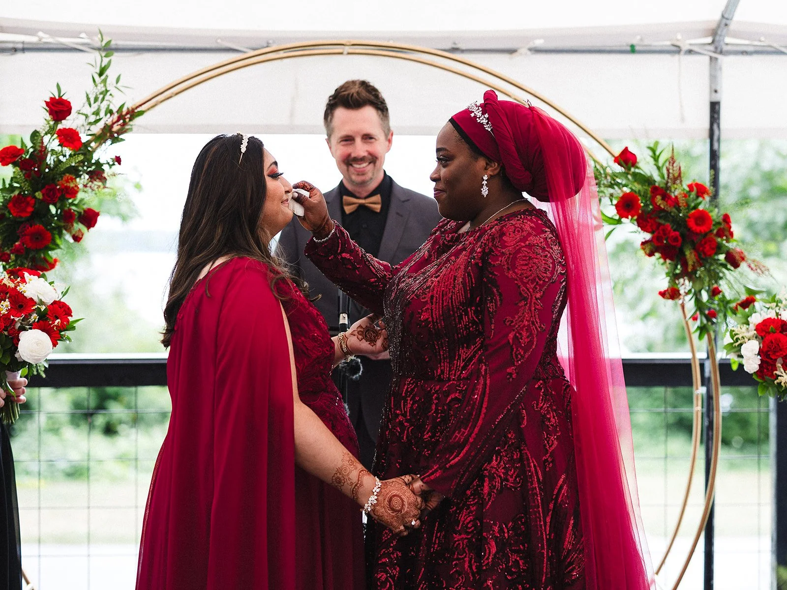 Brides in red dresses holding hands during a wedding ceremony, one bride is wiping a tear from the other's face, with an officiant in a tuxedo in the background and floral decorations around.