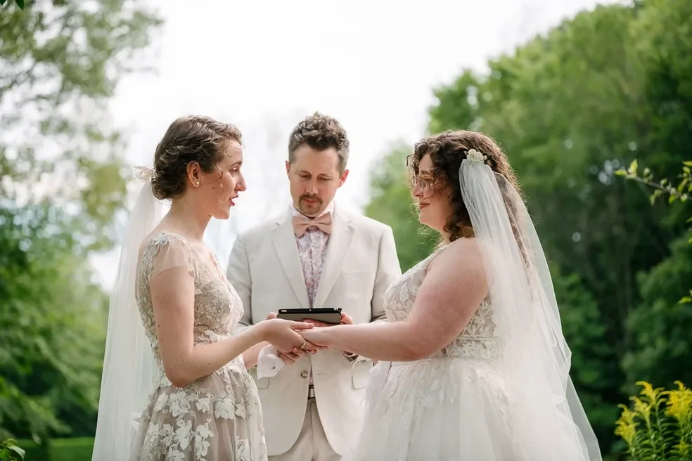 Two brides and one officiant during a wedding ceremony outdoors, with trees in the background.