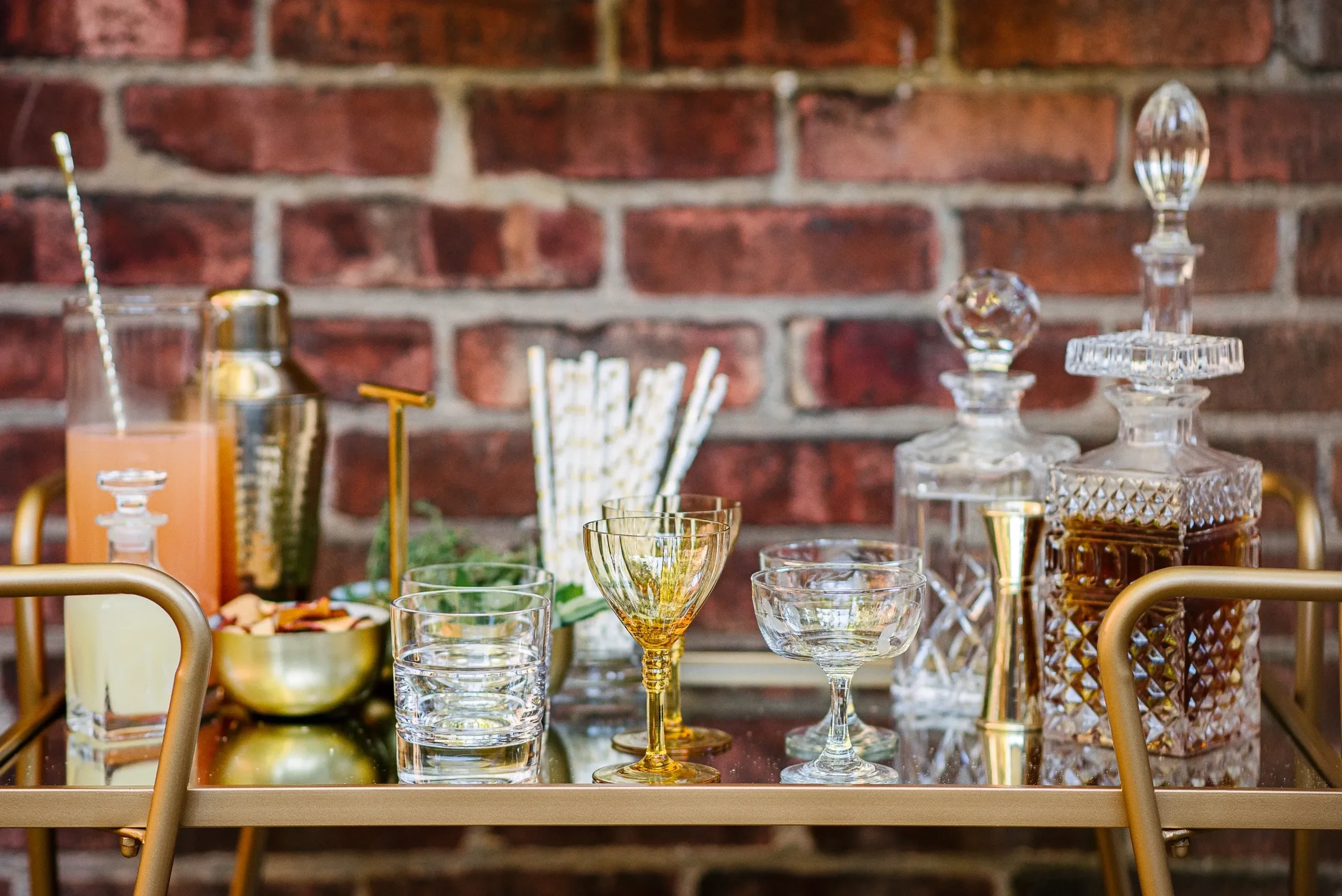 Bar cart with glasses, decanters, and drinks against a brick wall