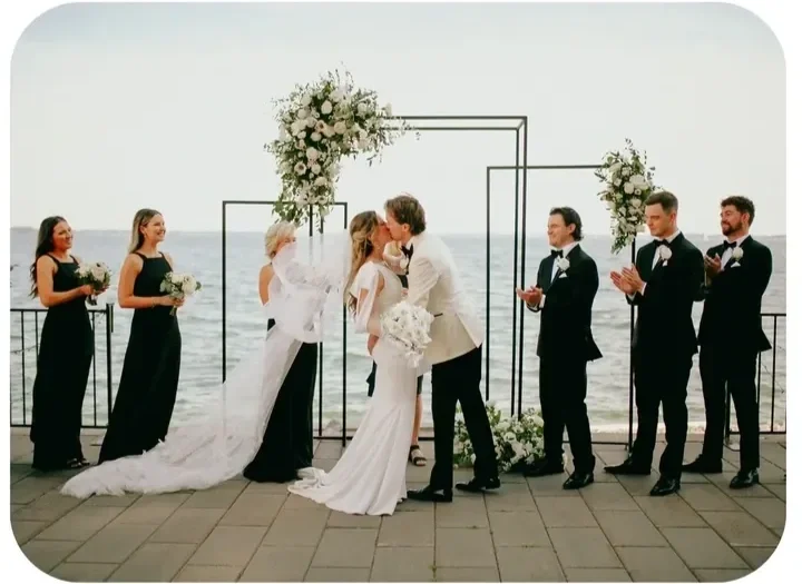 A wedding ceremony taking place outdoors by Lake Ontario, with the bride and groom kissing under a steel black archs, surrounded by bridesmaids and groomsmen in formal attire.