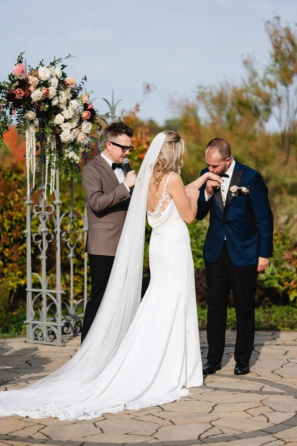 A bride and groom exchanging vows during an outdoor wedding ceremony, with a officiant reading from a microphone, in front of floral decorations and autumn trees.