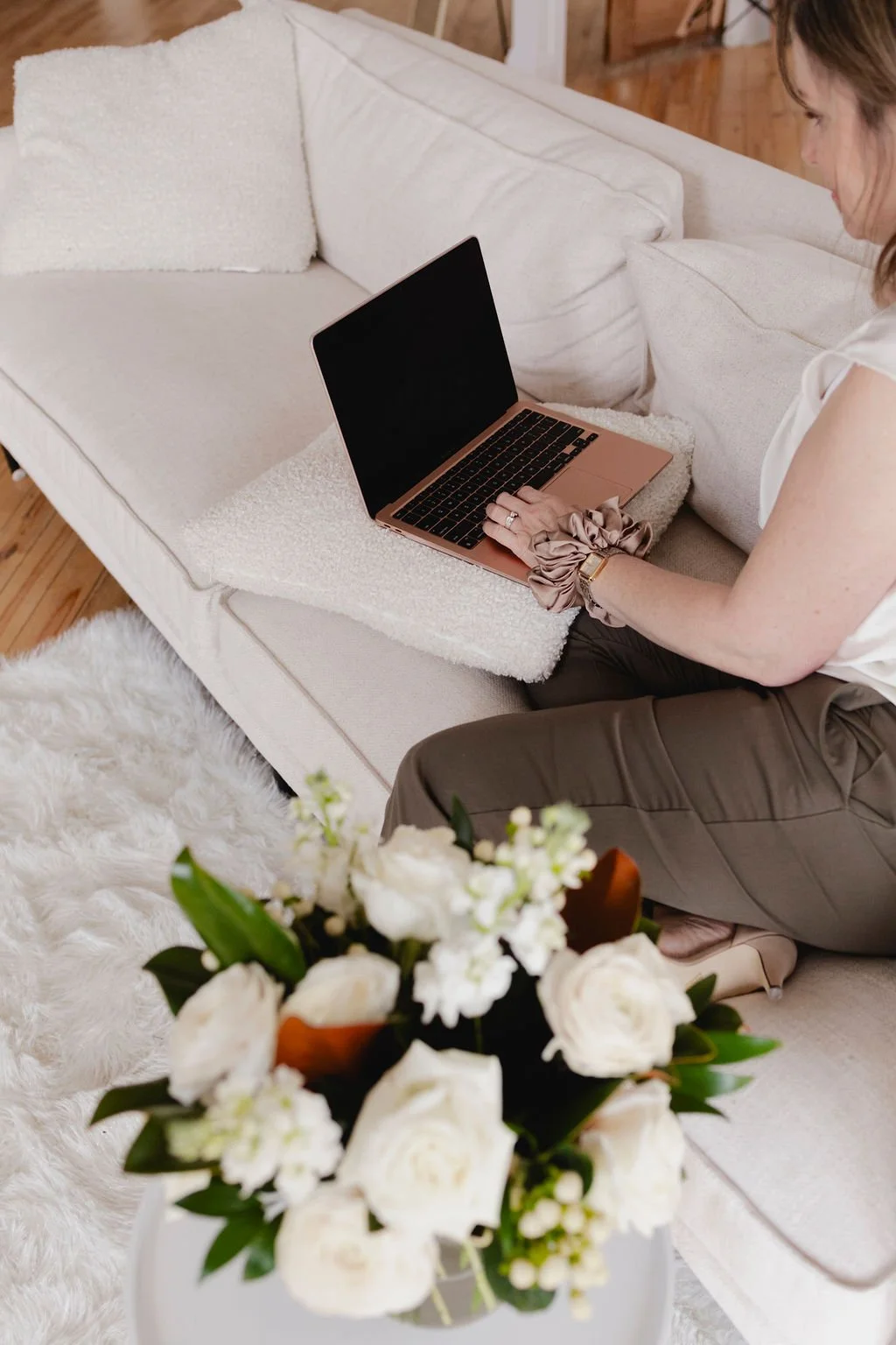 Wedding Planner sitting on a beige couch with a fluffy white blanket, using a pink laptop. A bouquet of white flowers is in the foreground.
