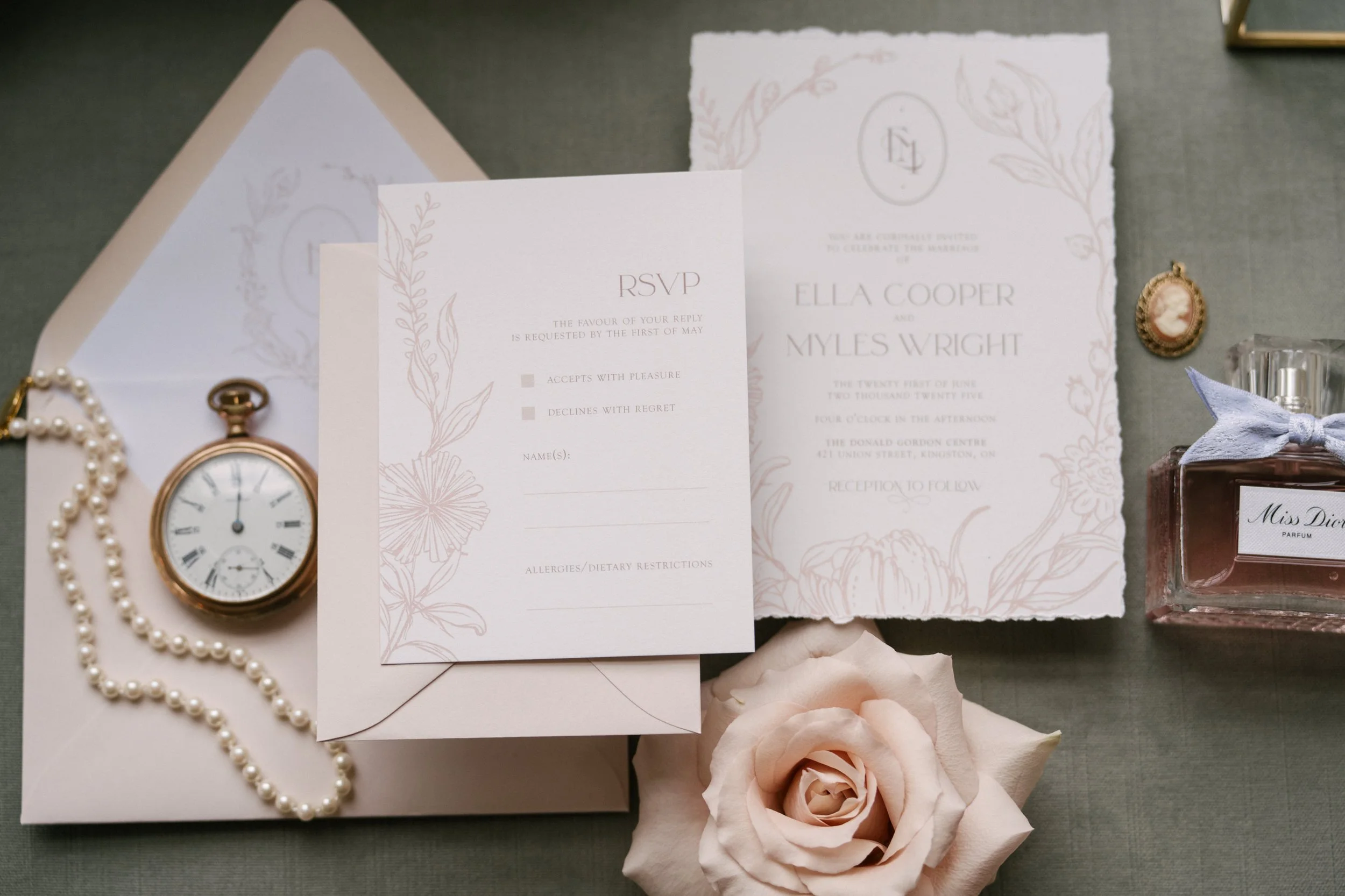Wedding invitation and RSVP card, clock, pearl necklace, pink rose, perfume bottle, cameo brooch, decorative papers on a gray surface.