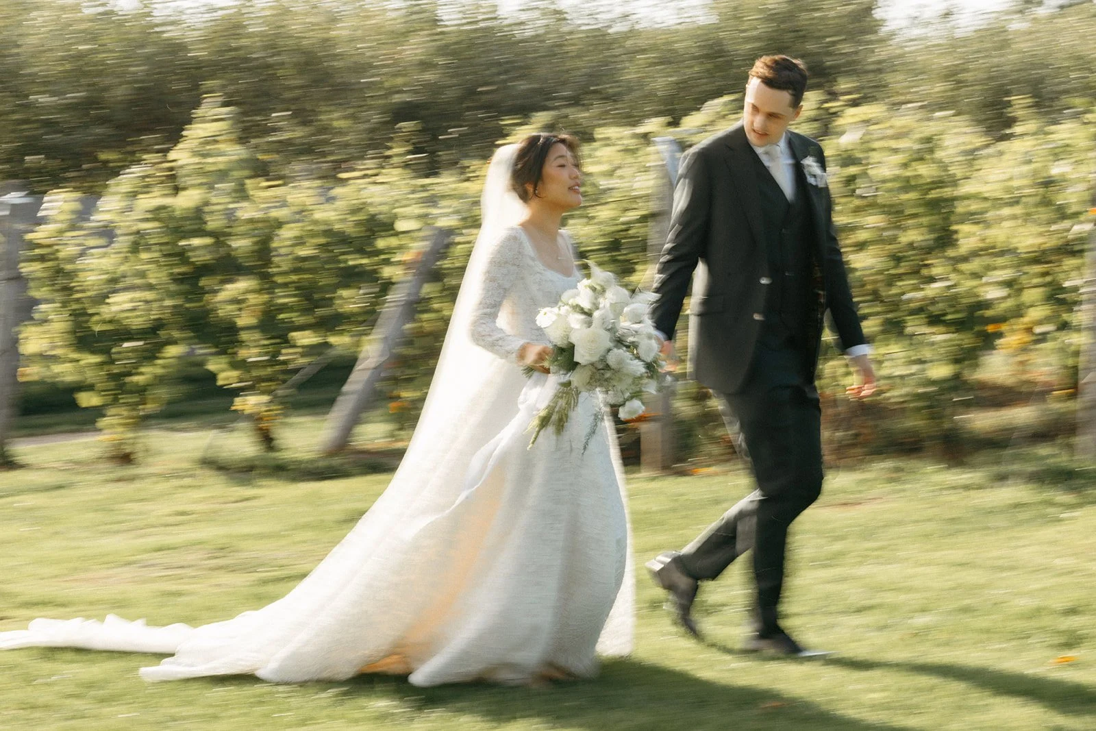 A bride and groom walking together outdoors through vineyards, the bride holding a large bouquet of white flowers.