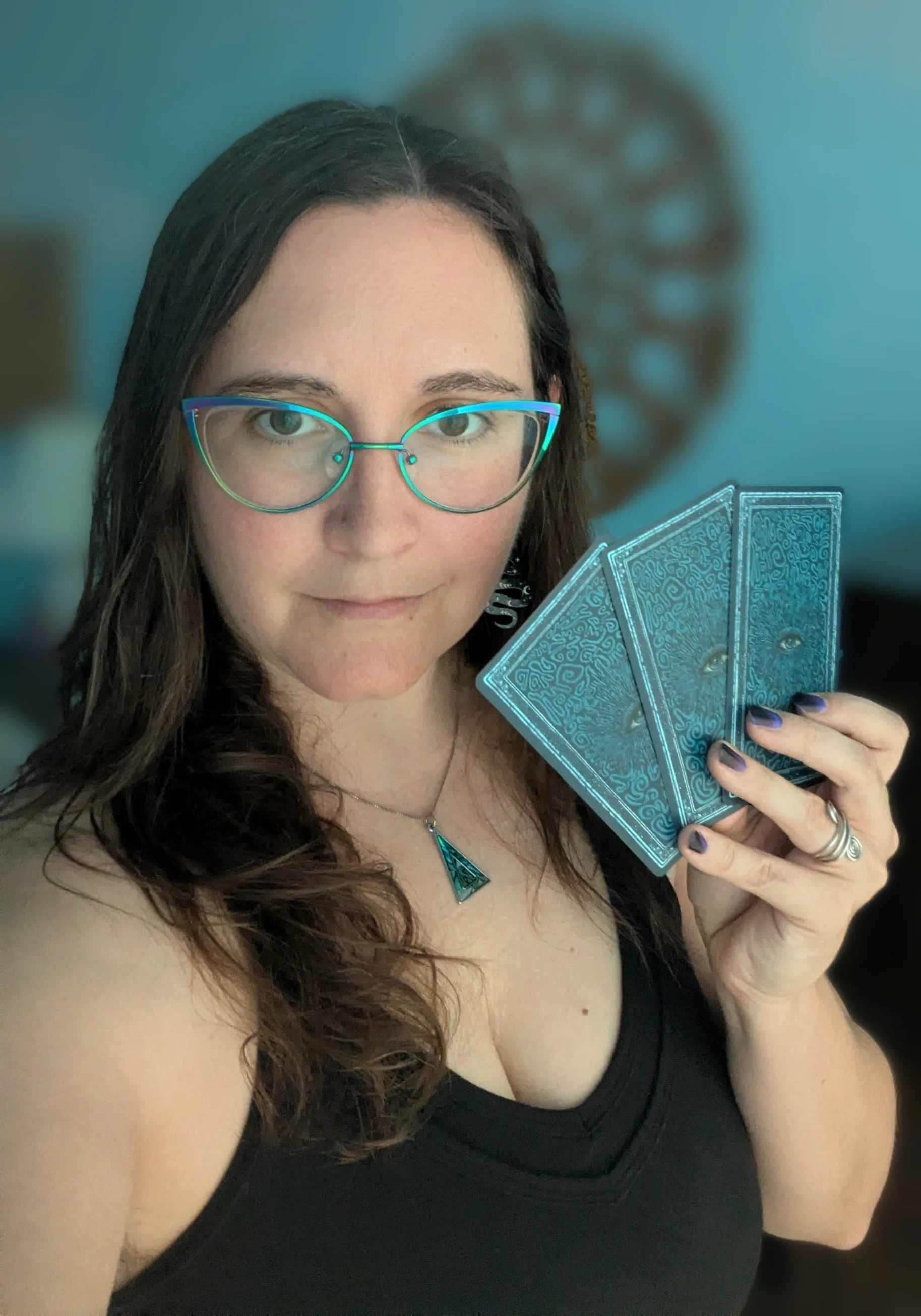 A woman with dark hair and glasses wearing a black top and holding a set of blue tarot cards.