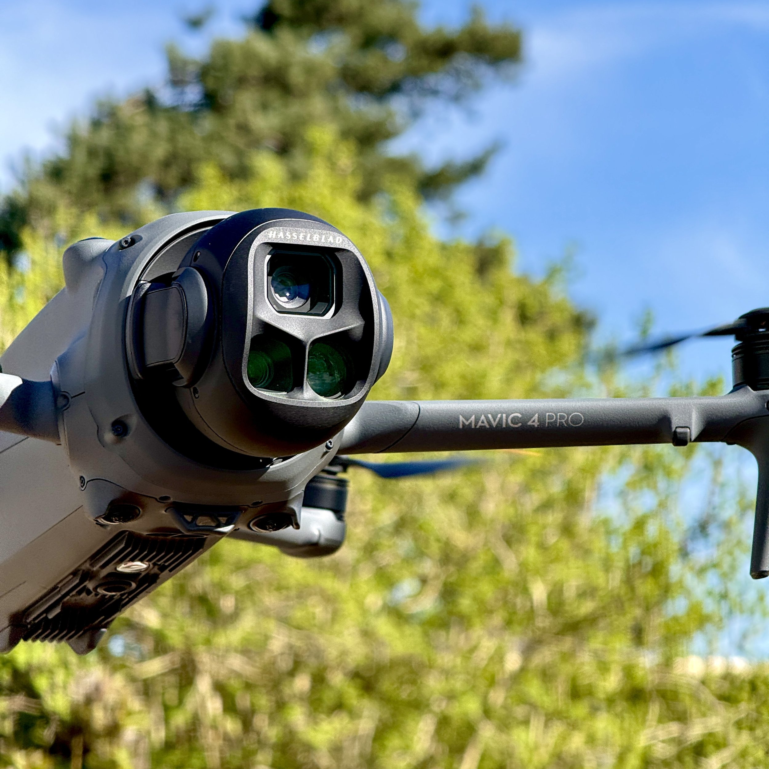 Close-up of a DJI Mavic 4 Pro drone with a Hasselblad camera lens, set against a background of green trees and blue sky.
