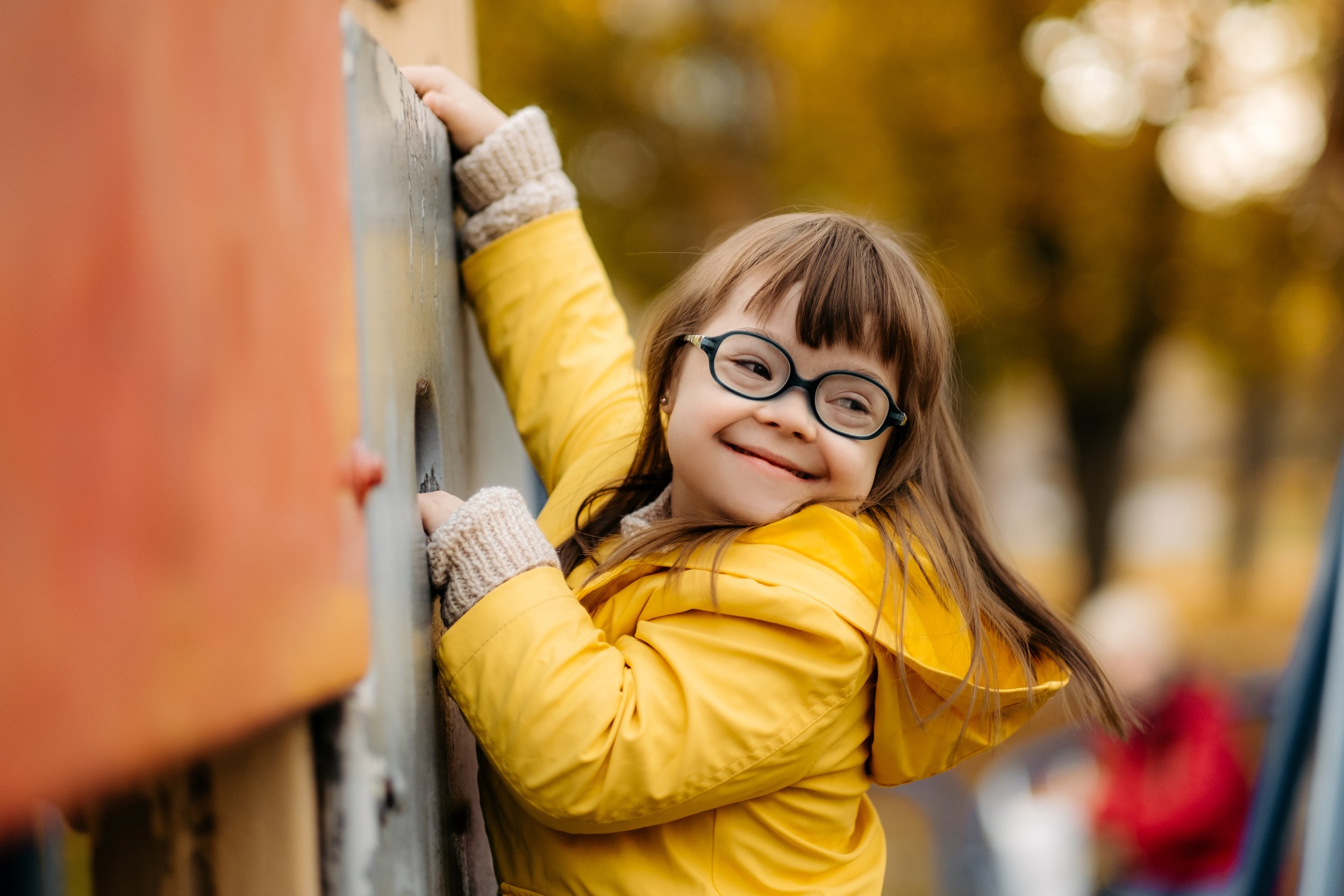 A young girl wearing glasses and a yellow jacket enjoys climbing on an outdoor structure.