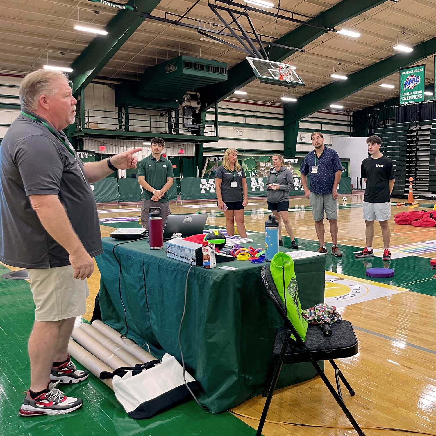 A coach giving instructions to a group of athletes in an indoor gymnasium with a basketball hoop with various sports equipment on a table. The athletes are standing and listening attentively.