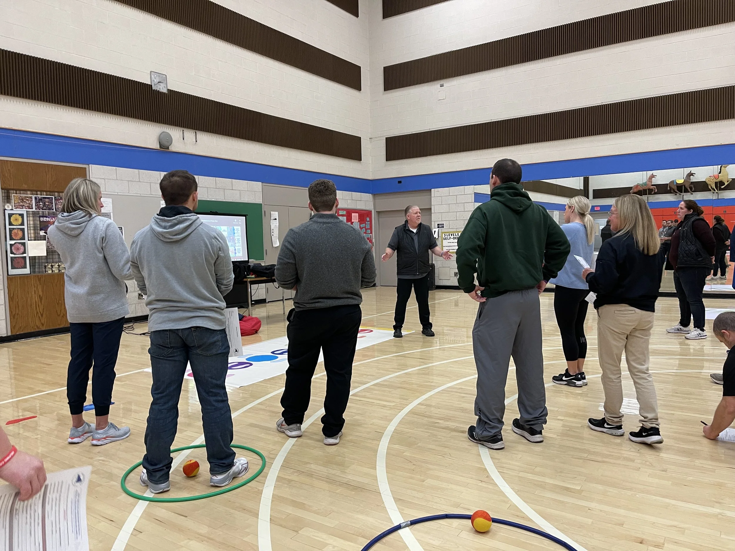 Group of people gathered in a gymnasium, listening to a man speaking. The gym has wooden floors and decorated walls with, ornaments, and a display with photographs. Some people are outdoors, standing near the wall, and there are hula hoops and soft balls on the floor.