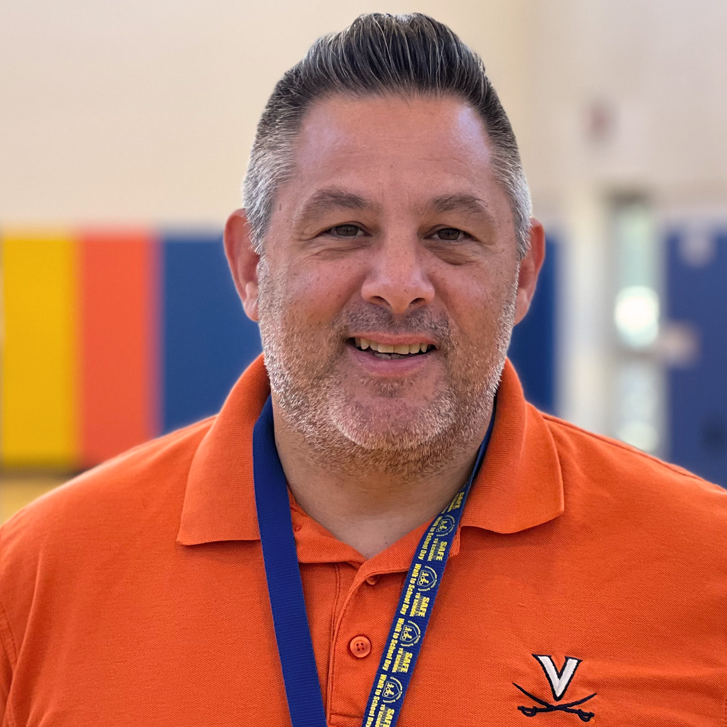 A middle-aged man smiling wearing an orange polo shirt with a Virginia Cavaliers logo, and a blue lanyard around his neck, standing indoors with blurred colorful lockers in the background.