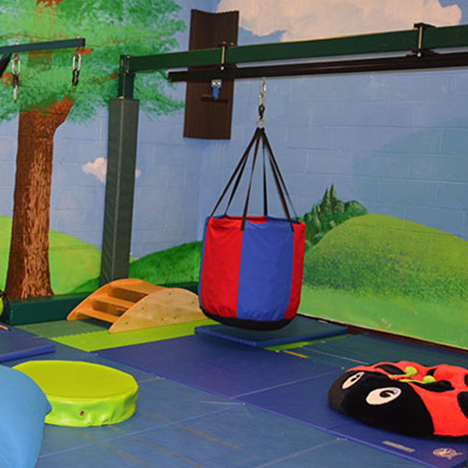 Indoor play area with a painted mural of trees and hills, featuring a climbing ramp, a hanging red and blue swing, and soft play mats on the floor.