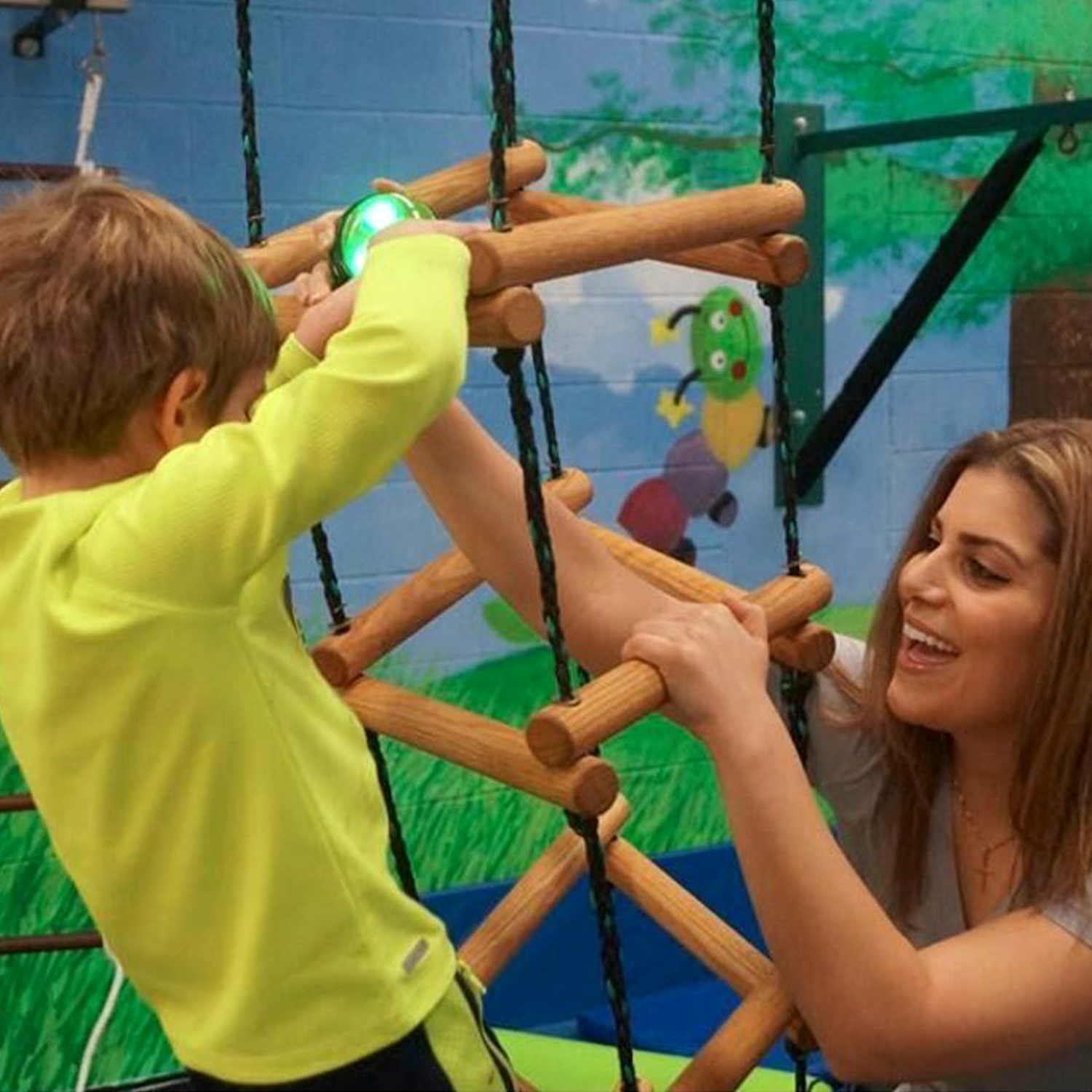 A young boy with short reddish hair in a yellow shirt climbing a wooden play structure while an adult woman helps and interacts with him in an indoor playground decorated like a forest scene.