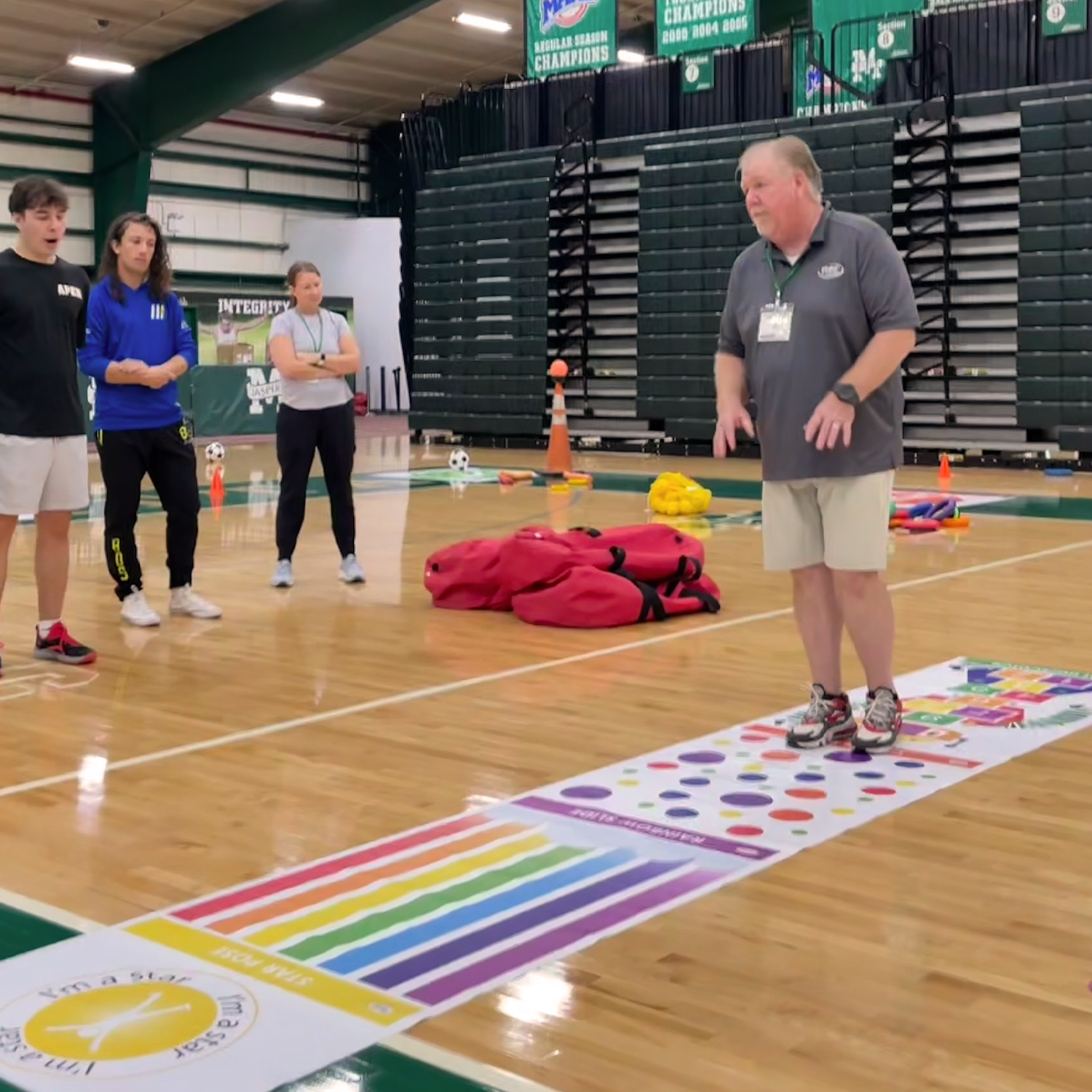 Group of people playing Twister on a gymnasium floor, with a coach or instructor standing nearby.