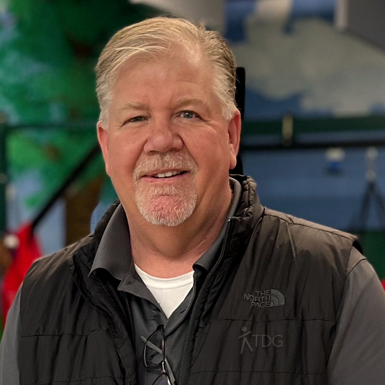 A smiling older man with gray hair and a goatee wearing a black vest and a gray shirt, sitting indoors with a blurred background.