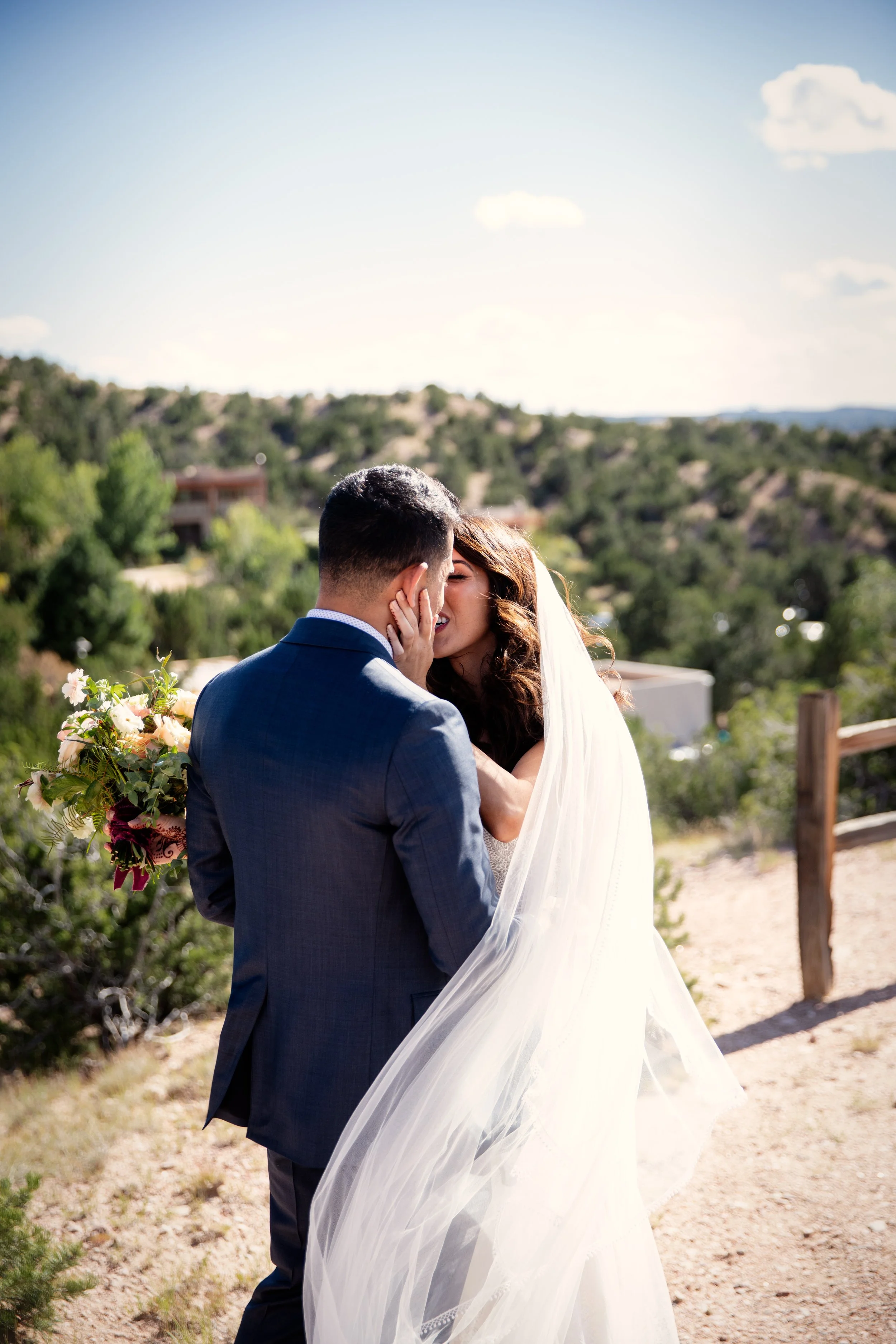 A bride and groom sharing a kiss outdoors on a sunny day, with the bride holding a bouquet of flowers and wearing a wedding gown with a veil, in a natural, scenic setting with hills and trees in the background.