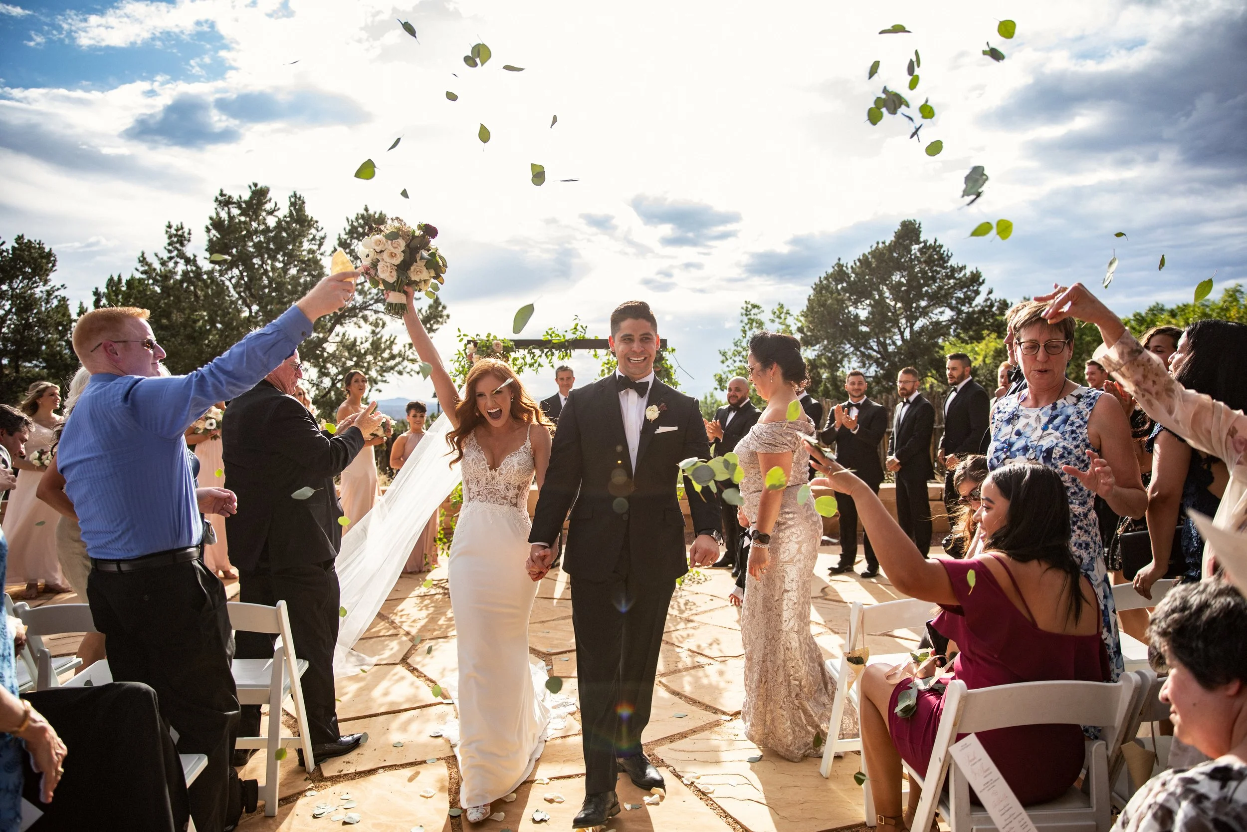 Wedding celebration with newlywed couple holding hands and walking down the aisle as guests throw flower petals and celebrate outdoors on a sunny day.