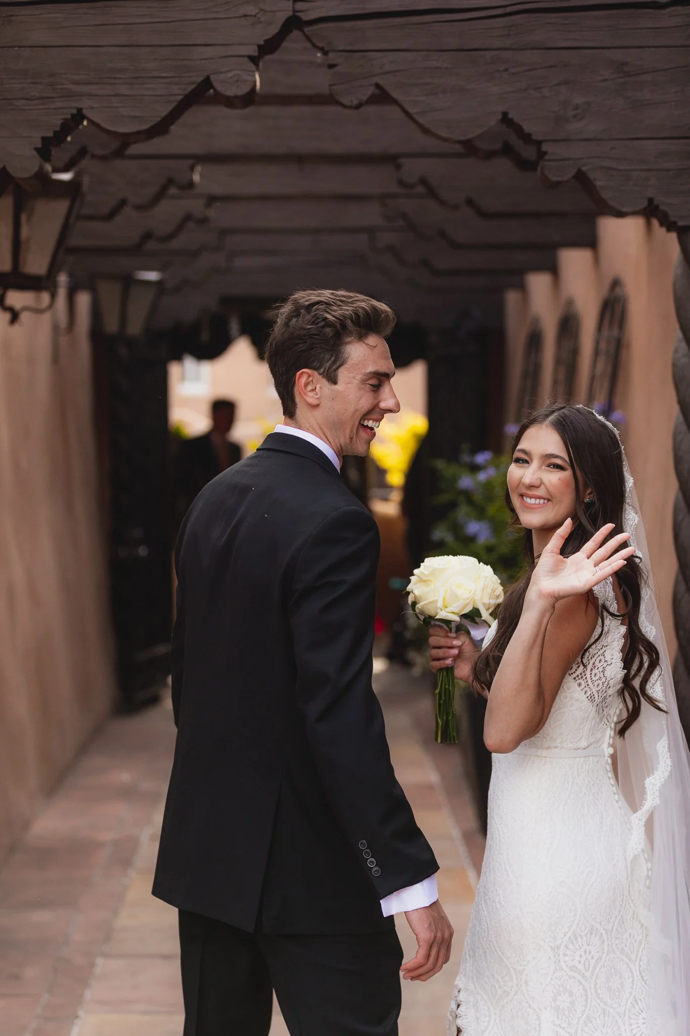 A bride and groom smiling at each other under a wooden archway, the bride holding a bouquet of white roses, celebrating their wedding.