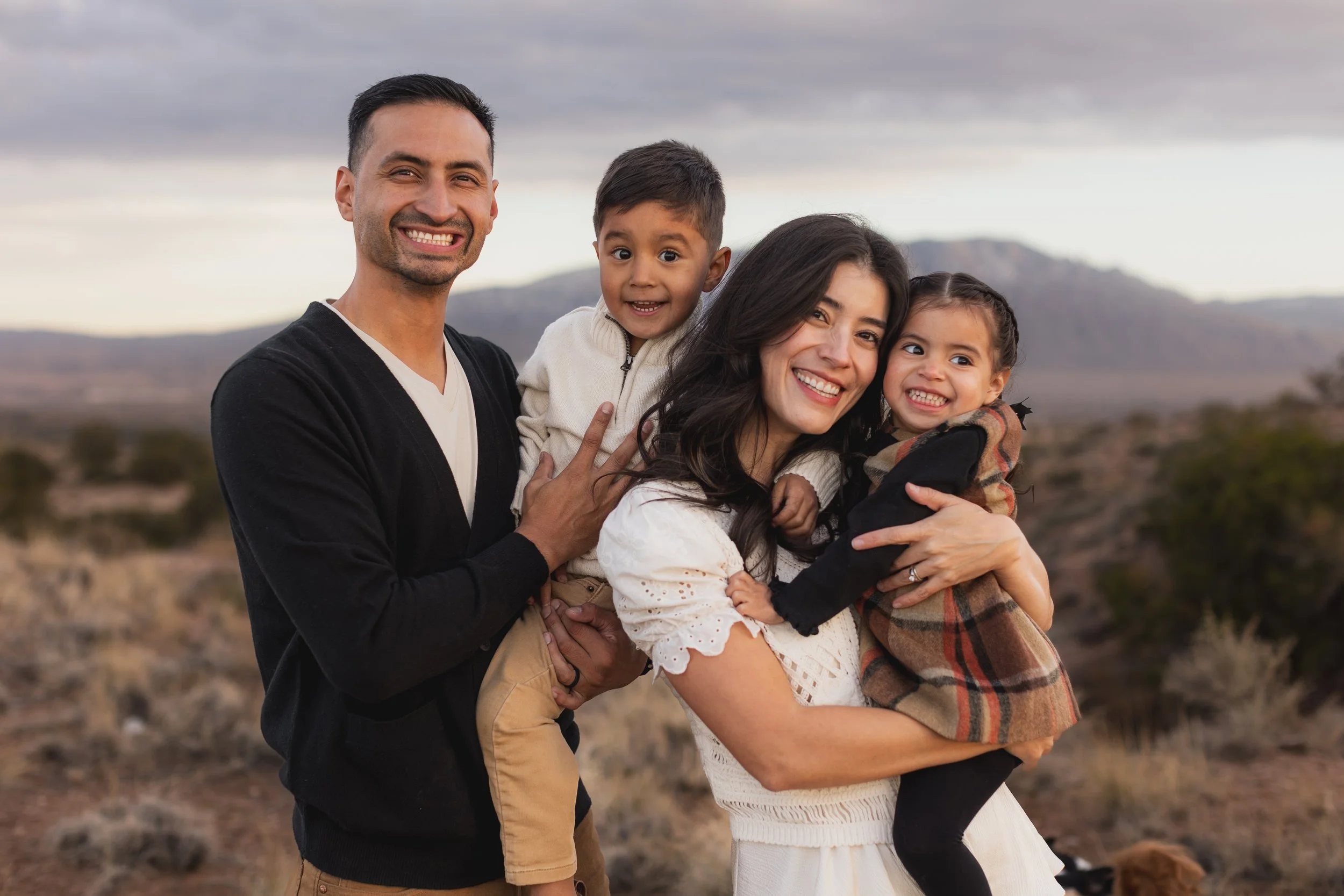 A family of four smiling outdoors in a desert landscape with mountains in the background.